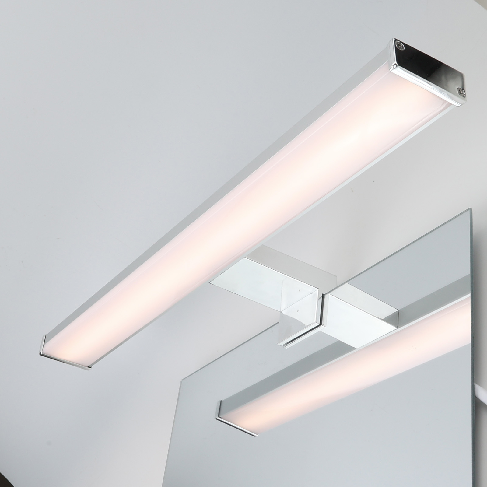 Una luz de tocador está montada encima de un espejo. La luz es plateada y rectangular, y el fondo es blanco.