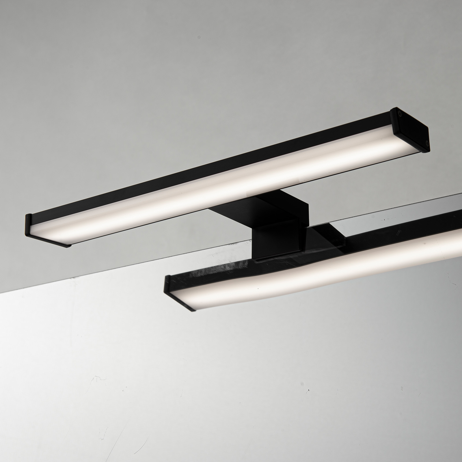Luminaria rectangular negra con dos barras paralelas iluminadas. Reflejada sobre una superficie gris claro. La luminaria tiene un diseño moderno.