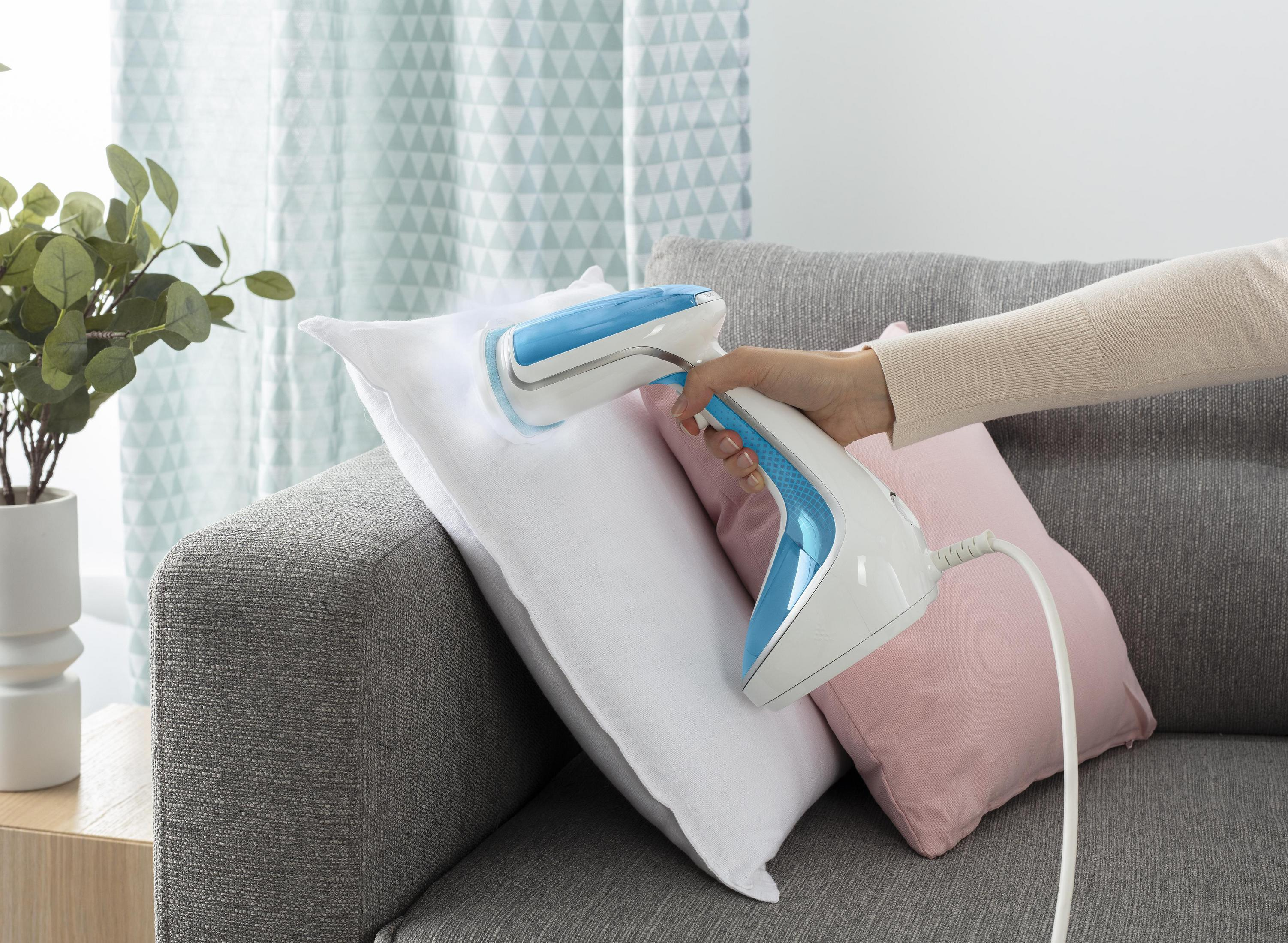 Ręka parująca białą poduszkę niebiesko-białym parownikiem. Szara sofa, różowa poduszka i roślina w tle.