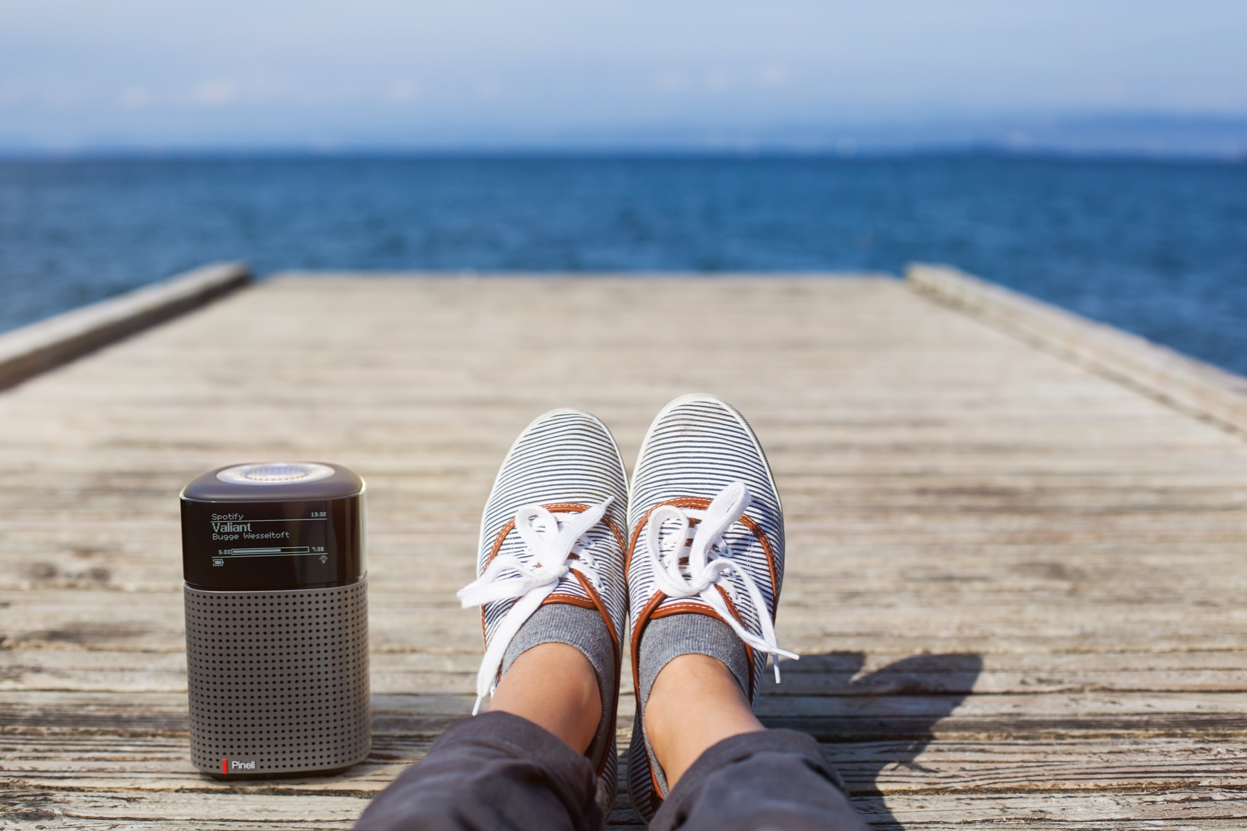Feet in striped sneakers resting on a wooden dock. A small radio speaker sits nearby. The sea and sky form the background.