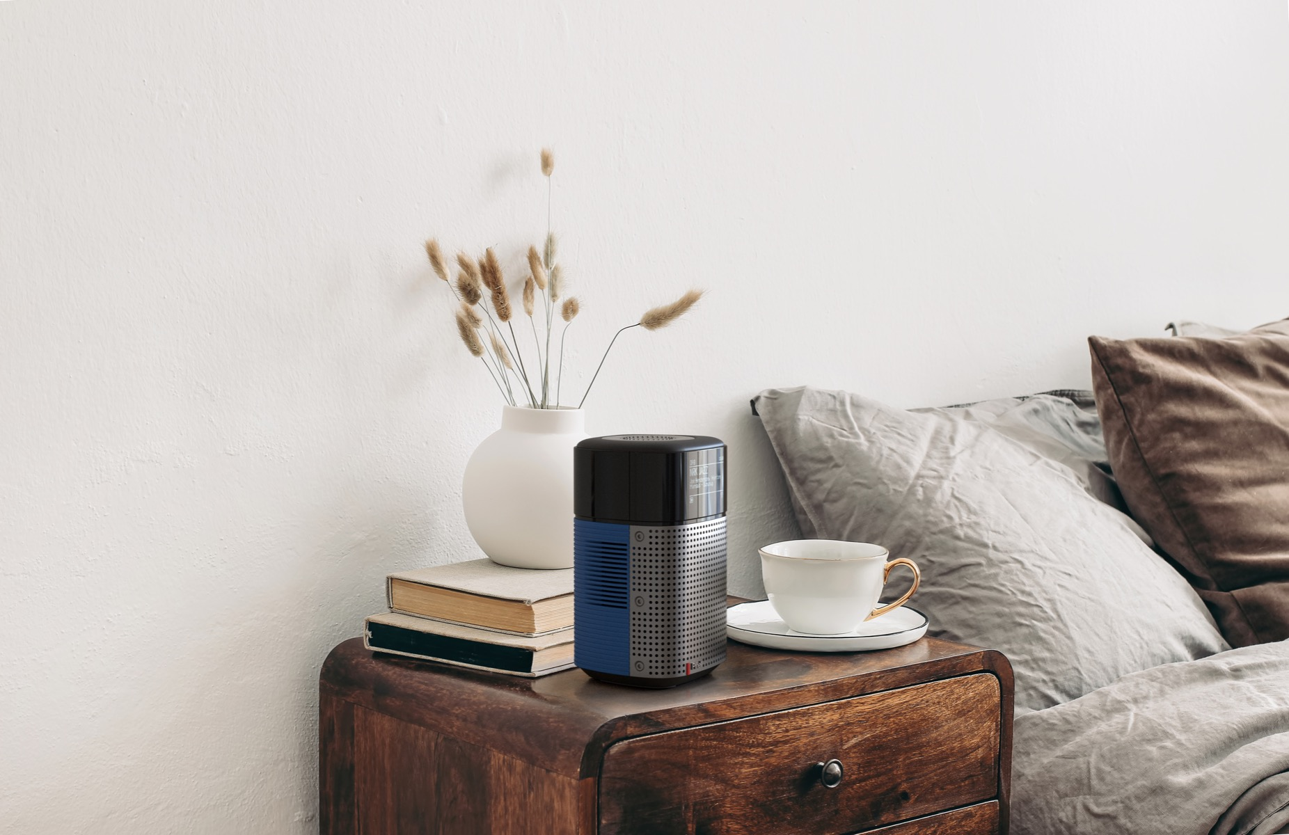 A bedside table with a vase, books, and a speaker. A cup and saucer sit next to the speaker. A bed is in the background.