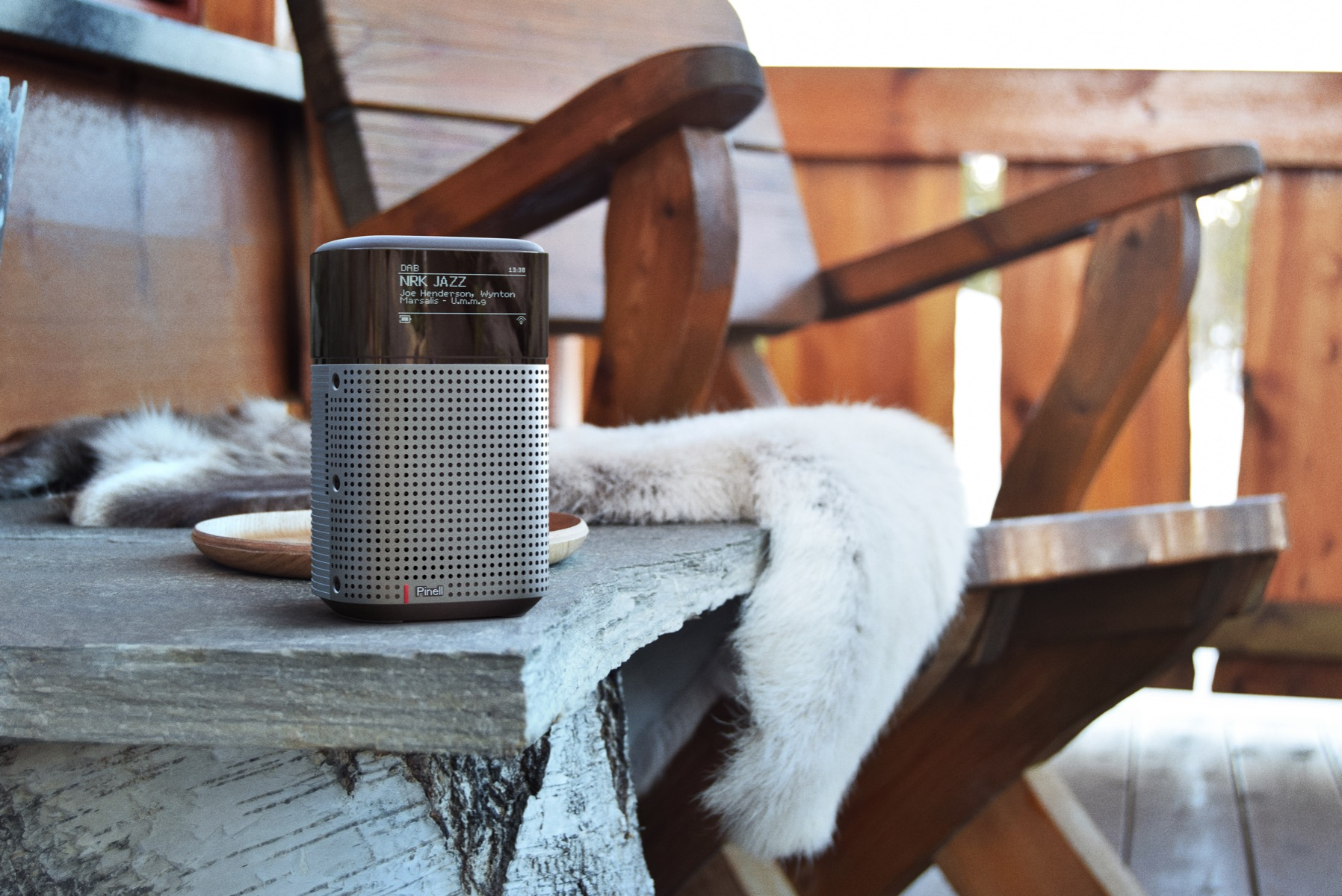 A speaker is on a table with a chair, a fur, and a wooden bowl.
