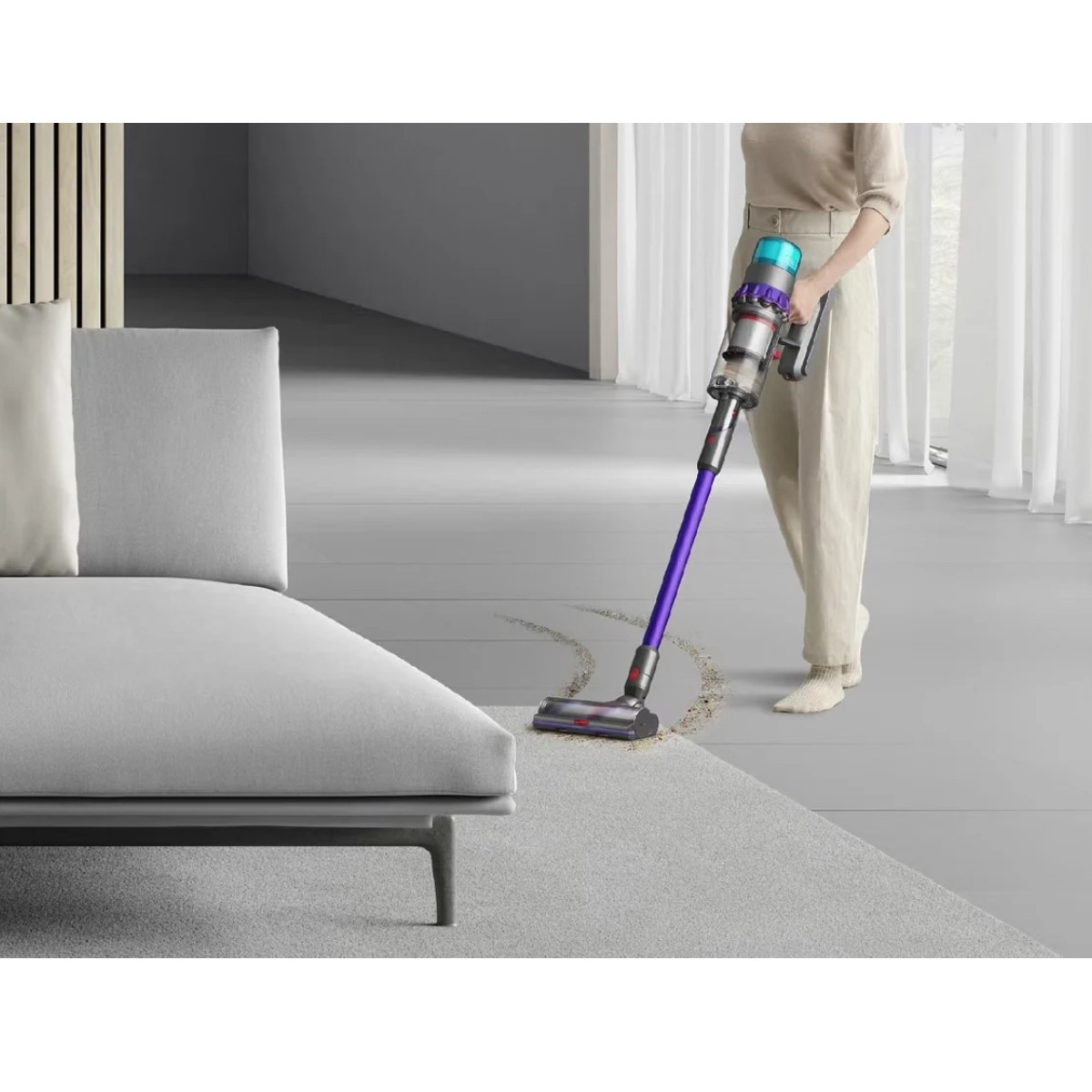 Osoba odkurza dywan fioletowym i srebrnym odkurzaczem. Szara kanapa po lewej, okno z zasłonami w tle.
