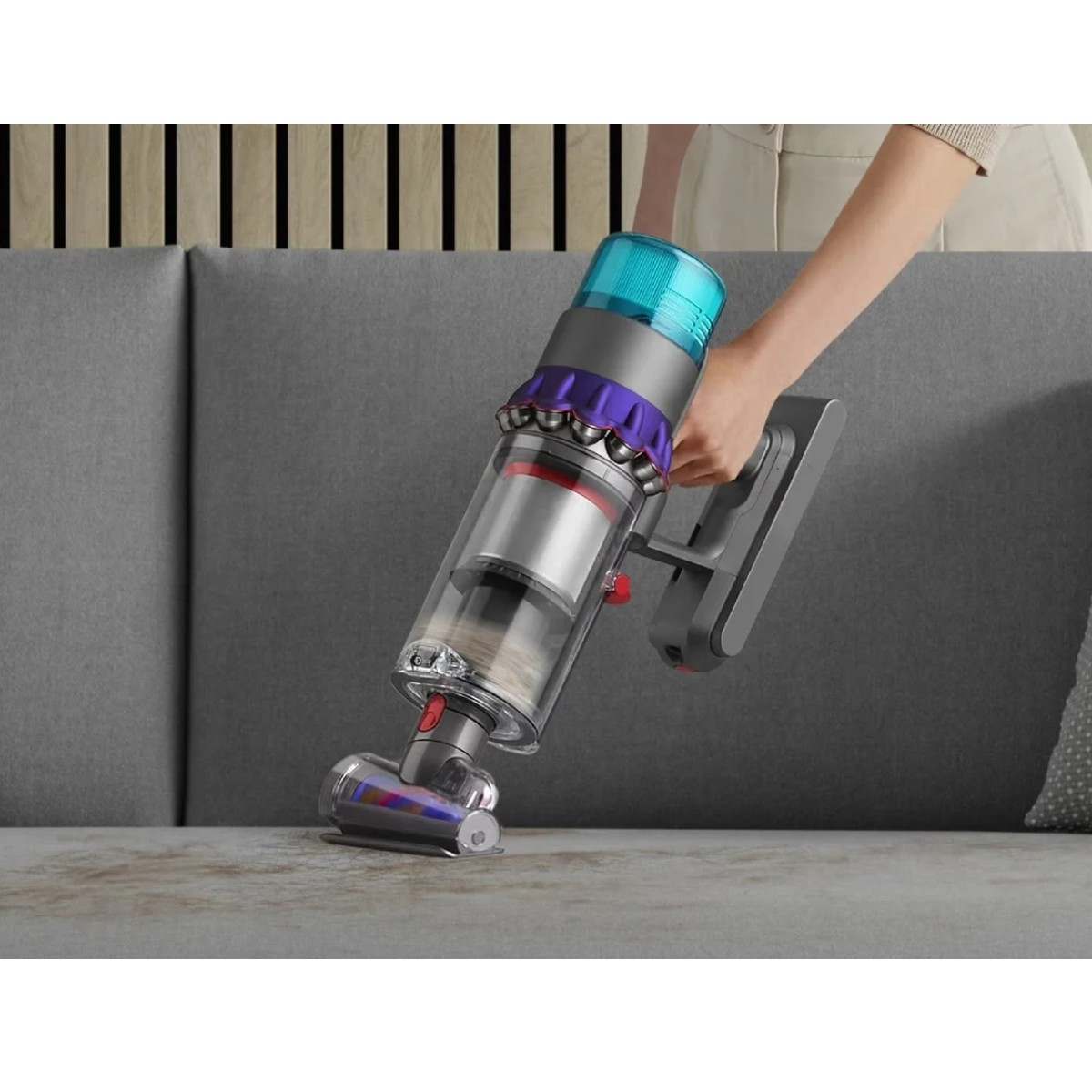 Osoba odkurza kanapę fioletowym i srebrnym odkurzaczem. Kurz jest wciągany z kanapy do przezroczystego pojemnika.