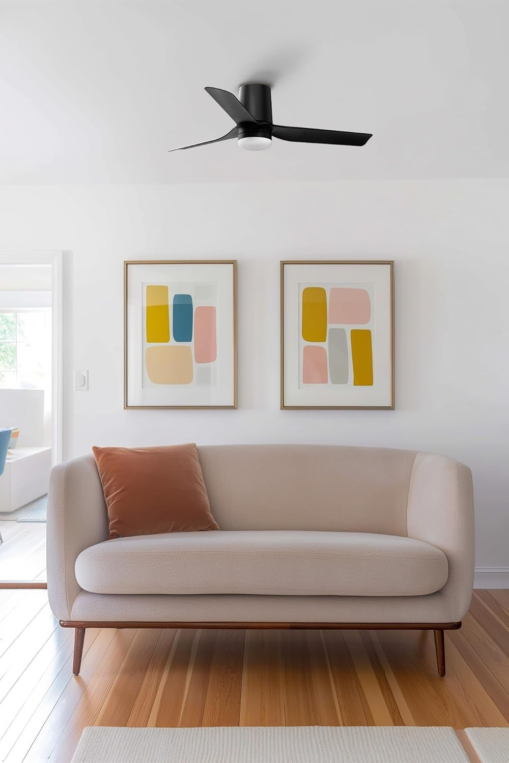 Soggiorno con divano beige, ventilatore a soffitto, due opere d'arte incorniciate, un cuscino arancione e pavimento in legno.