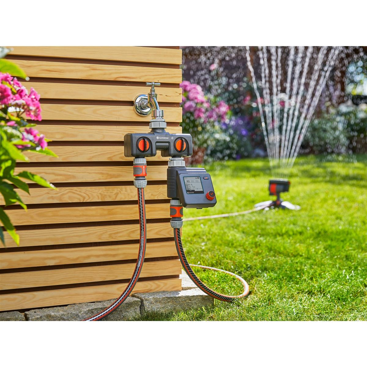 Ein Gartenschlauchsystem mit Zeitschaltuhr ist an einen Wasserhahn angeschlossen. Ein Rasensprenger sprüht Wasser auf einen grünen Rasen.
