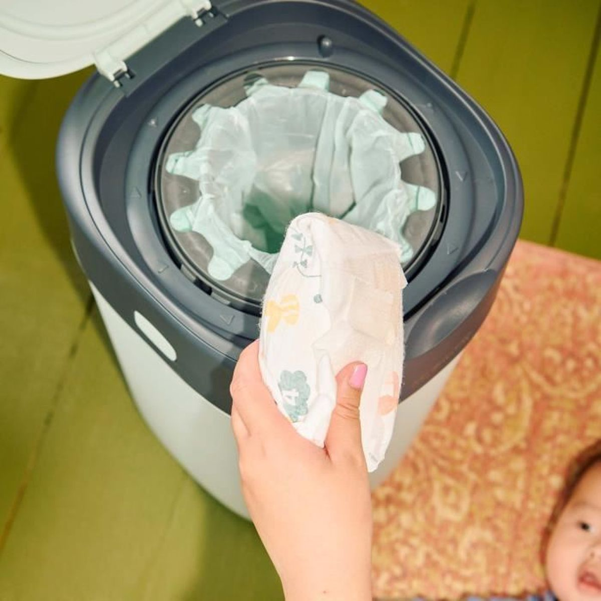 Eine Hand hält eine Windel über einem Windelentsorgungssystem. Das Gerät ist geöffnet, mit einem vollen Beutel im Inneren, und ein Baby ist im Hintergrund.