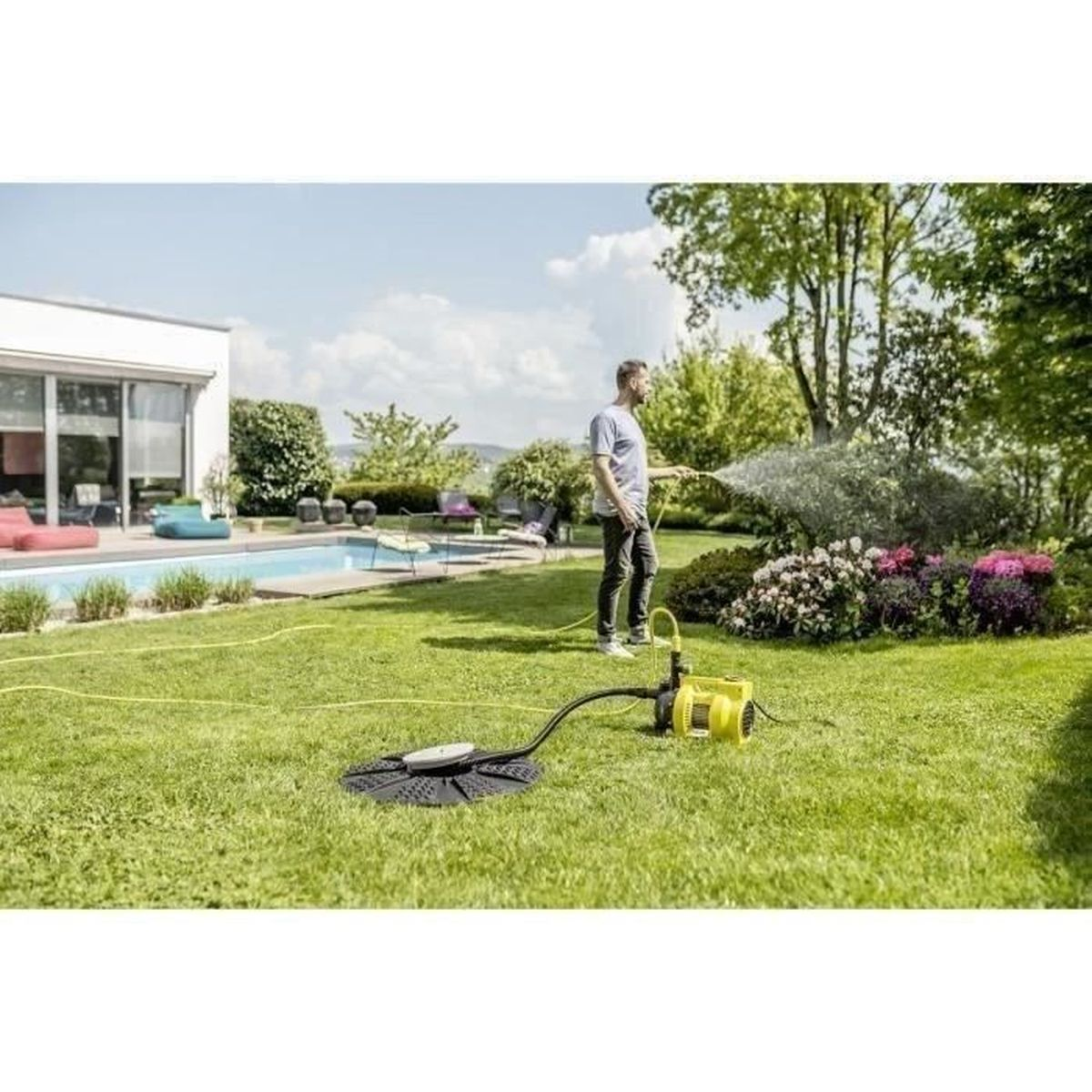 Ein Mann bewässert Pflanzen in einem Garten mit einer gelben Pumpe und einem Schlauch, umgeben von grünem Gras und Bäumen.