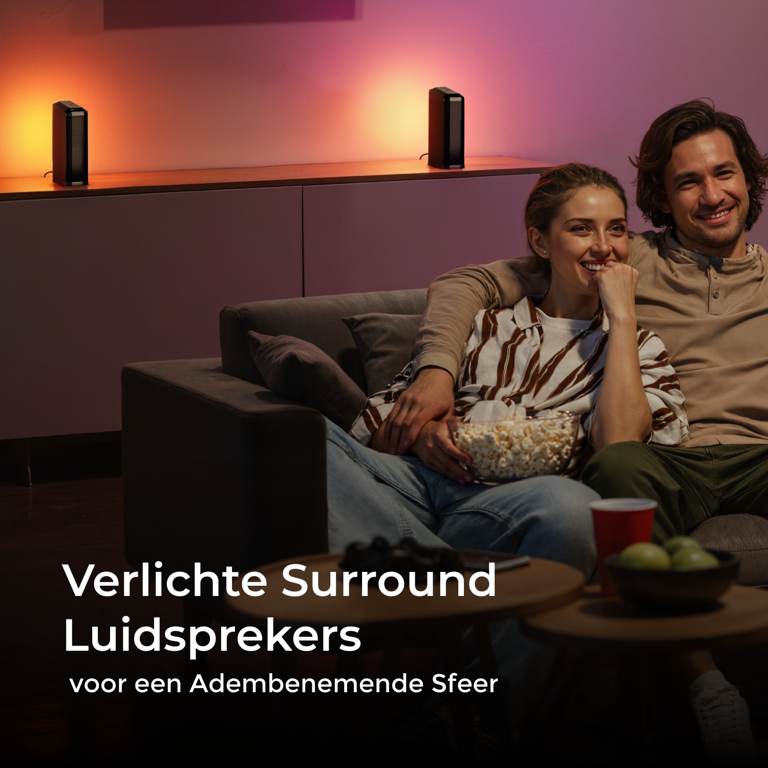 A couple sitting on a sofa watching TV with speakers and popcorn.