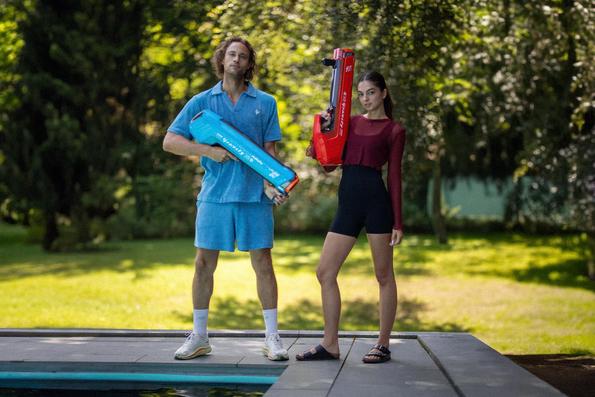 Zwei Personen stehen an einem Pool, eine hält eine Wasserpistole, die andere einen roten Wasserbombenwerfer, in einem Garten.