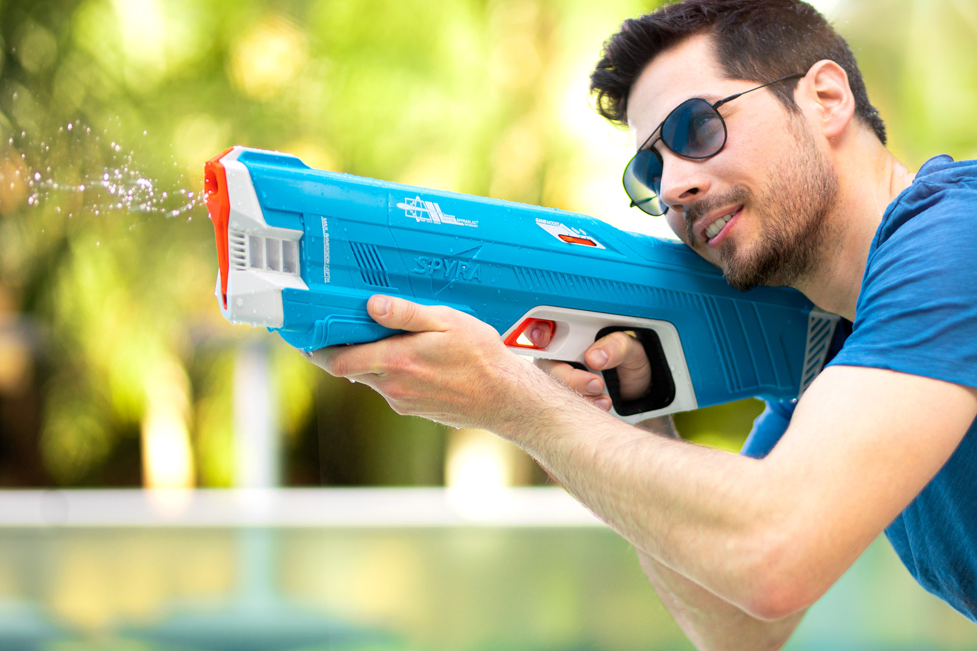 Ein Mann mit Sonnenbrille zielt mit einer blauen Wasserpistole und sprüht Wasser. Der Hintergrund ist verschwommenes grünes Laub.
