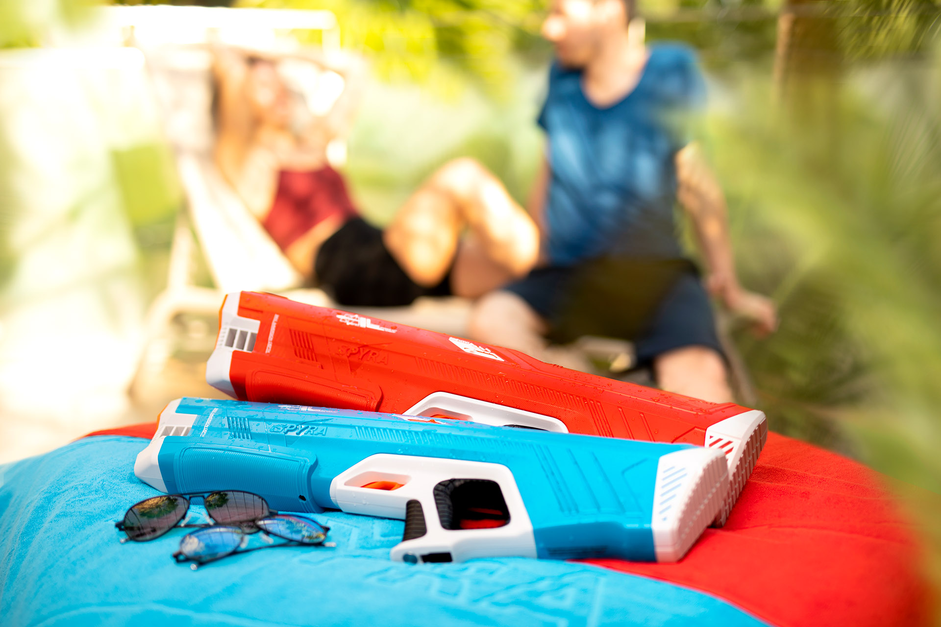 Zwei Wasserpistolen auf einem blauen Handtuch mit Sonnenbrillen. Zwei Personen sind im Hintergrund unscharf.