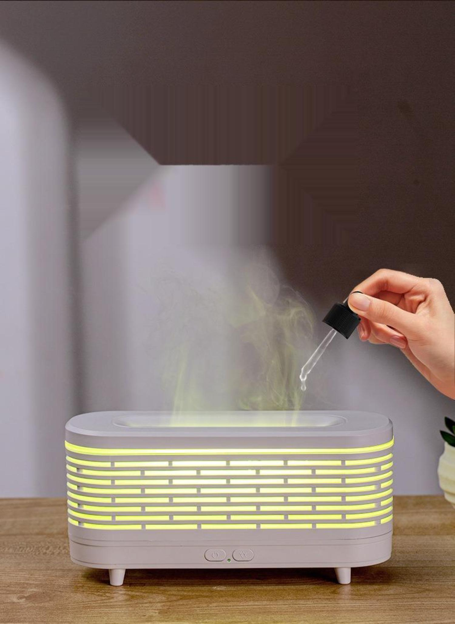 Ein Aromadiffusor mit Flammeneffekt. Es ist weiß und hat eine Entlüftung. Eine Hand gibt Öl hinzu.