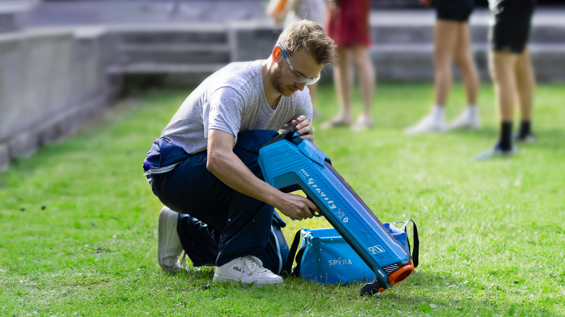 Ein Mann kniet auf Gras und baut eine blaue Wasserpistole zusammen. Andere Personen unscharf.