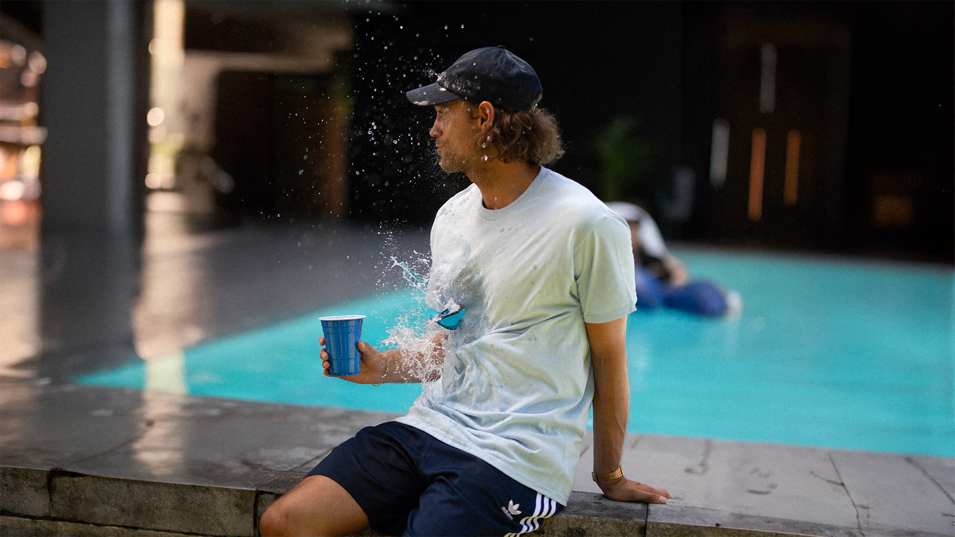 Mann in hellblauem Hemd, schwarzer Kappe und Shorts wird von Wasser getroffen.