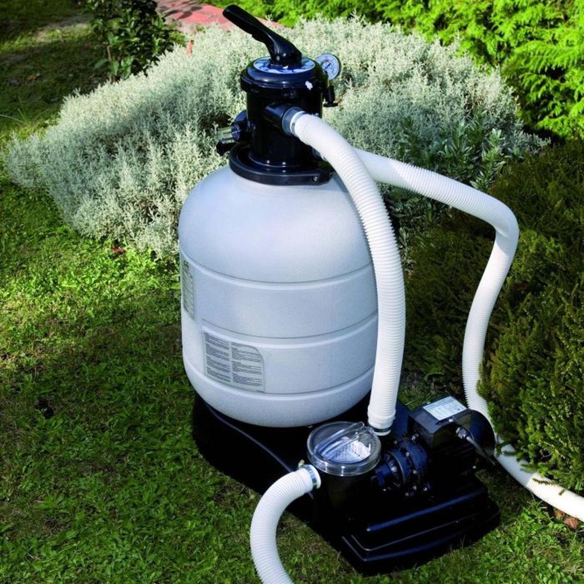 Un sistema de filtro y bomba de piscina sobre césped verde, con mangueras blancas y plantas alrededor.