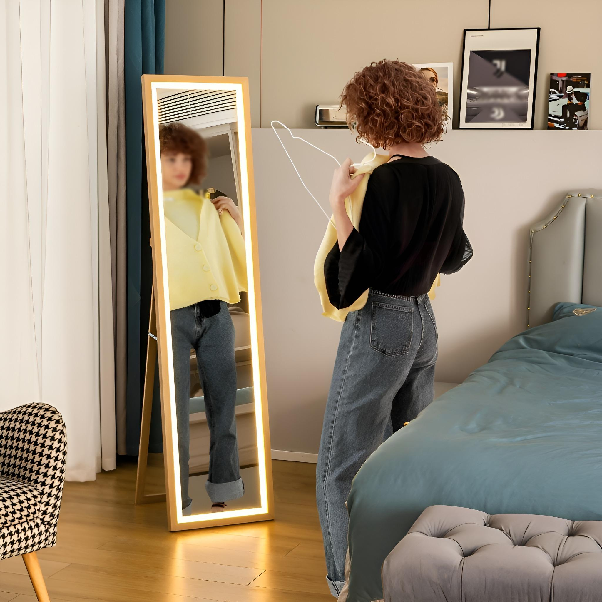 Frau in einem Schlafzimmer mit Spiegel. Sie hält einen gelben Pullover und spiegelt sich in dem beleuchteten Ganzkörperspiegel.