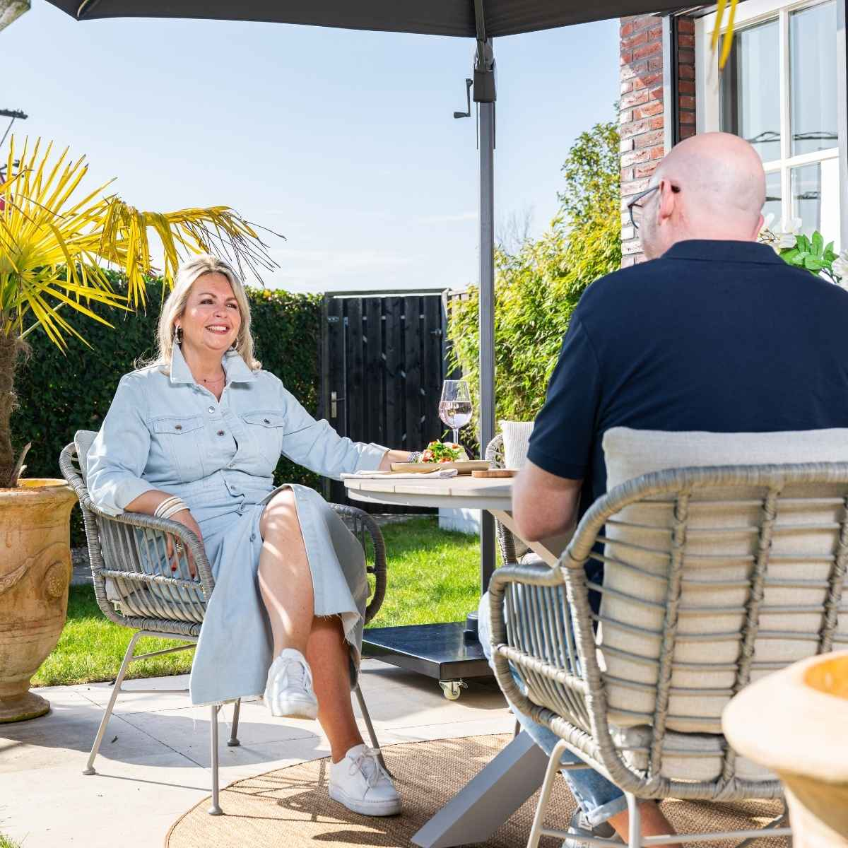 Een vrouw en een man die buiten aan een tafel zitten. Er is een parasol en een groen gazon.