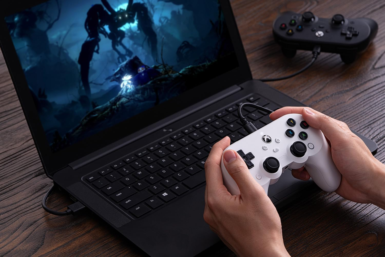 Dłonie trzymające biały kontroler gier, grające w grę na laptopie. Widoczny jest drugi czarny kontroler.
