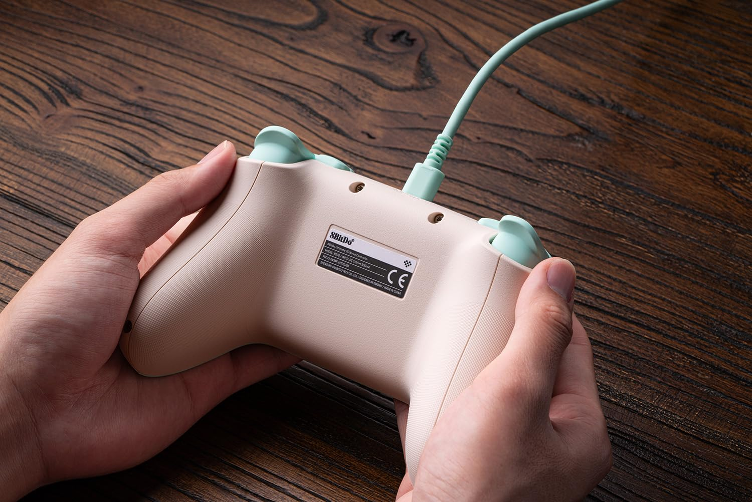 Dłonie trzymające tył jasno brązowego i zielonego kontrolera do gier z dołączonym zielonym przewodem.