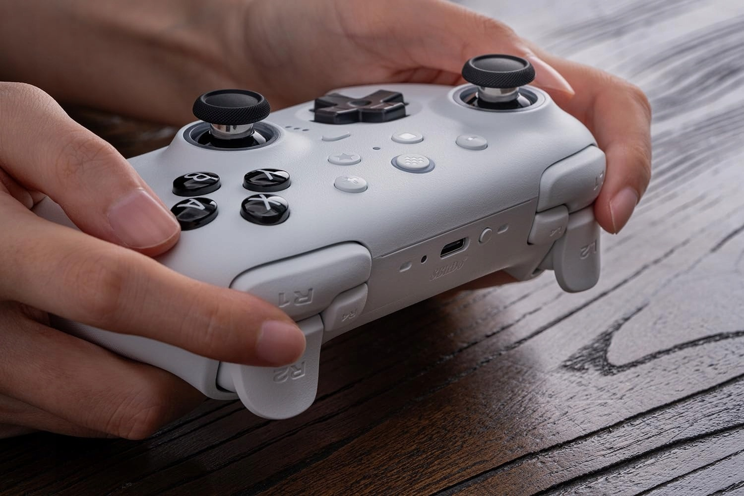 Ręce trzymające biały kontroler do gier z czarnymi joystickami i przyciskami.