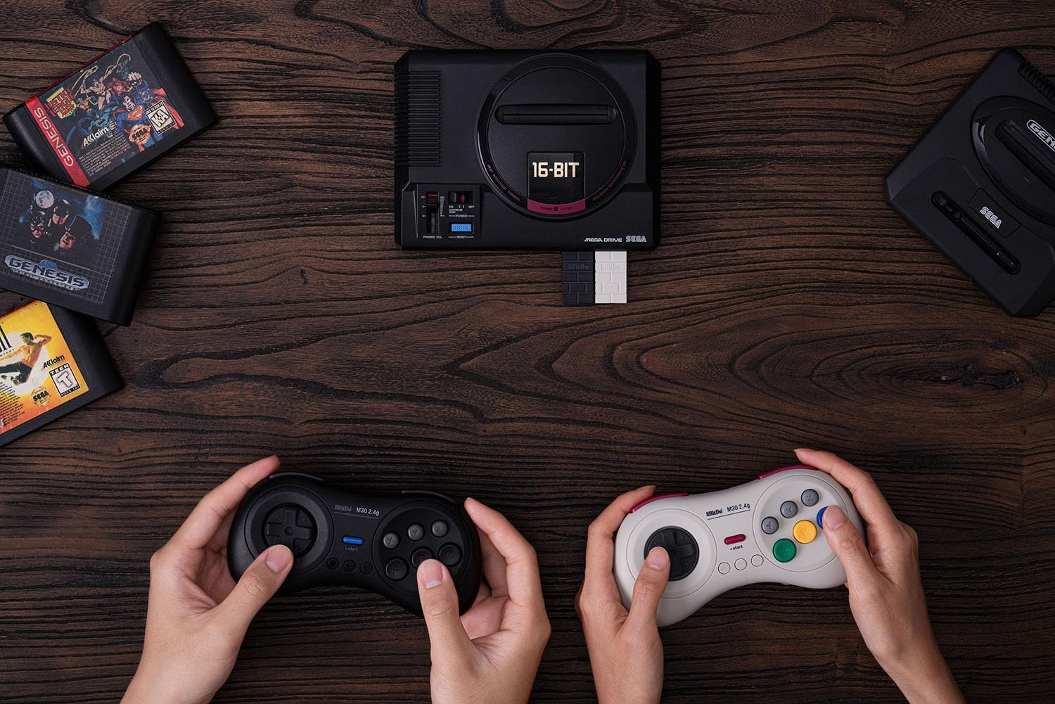Due mani tengono controller. Due console Sega Genesis e cartucce su un tavolo di legno.