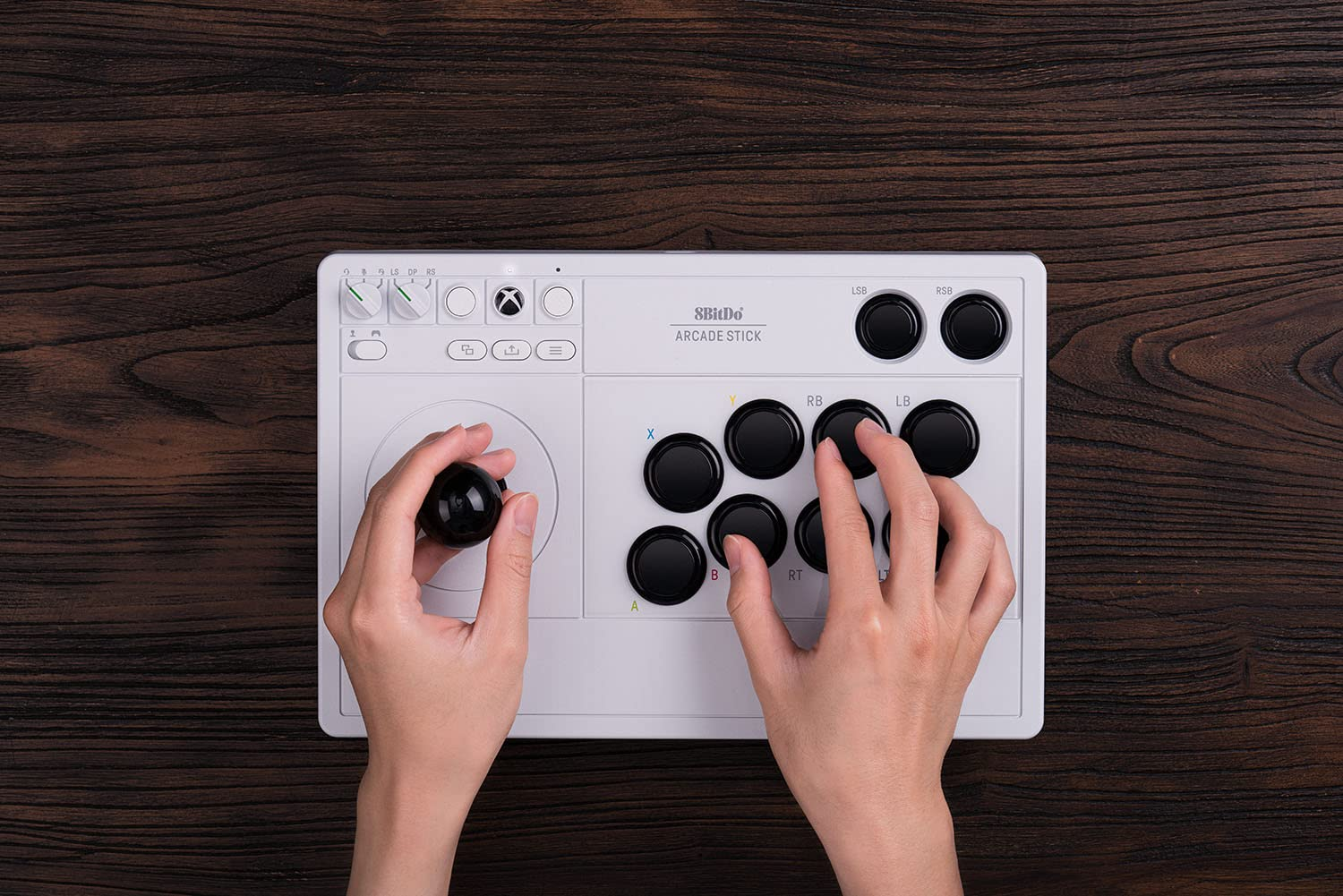 Dłonie na białym joysticku arcade. Joystick znajduje się na drewnianej powierzchni. Widoczne są przyciski i joystick.