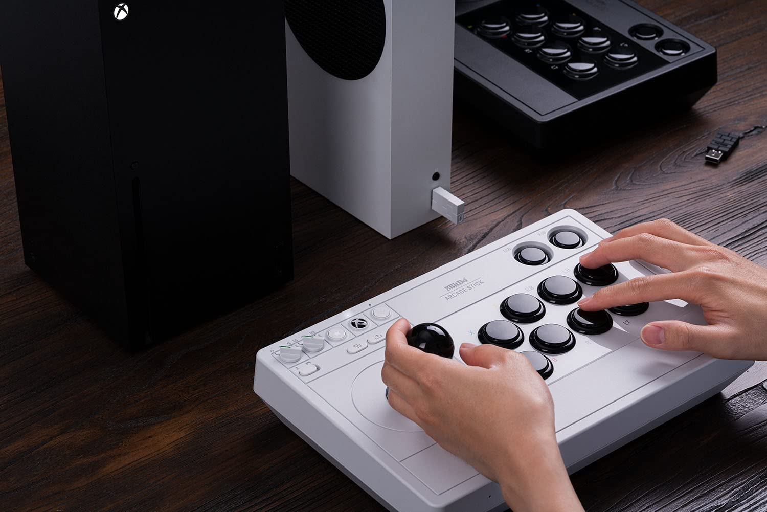 Joystick arcade obok dwóch konsol Xbox. Ręka jest na joysticku, a joystick ma przyciski.