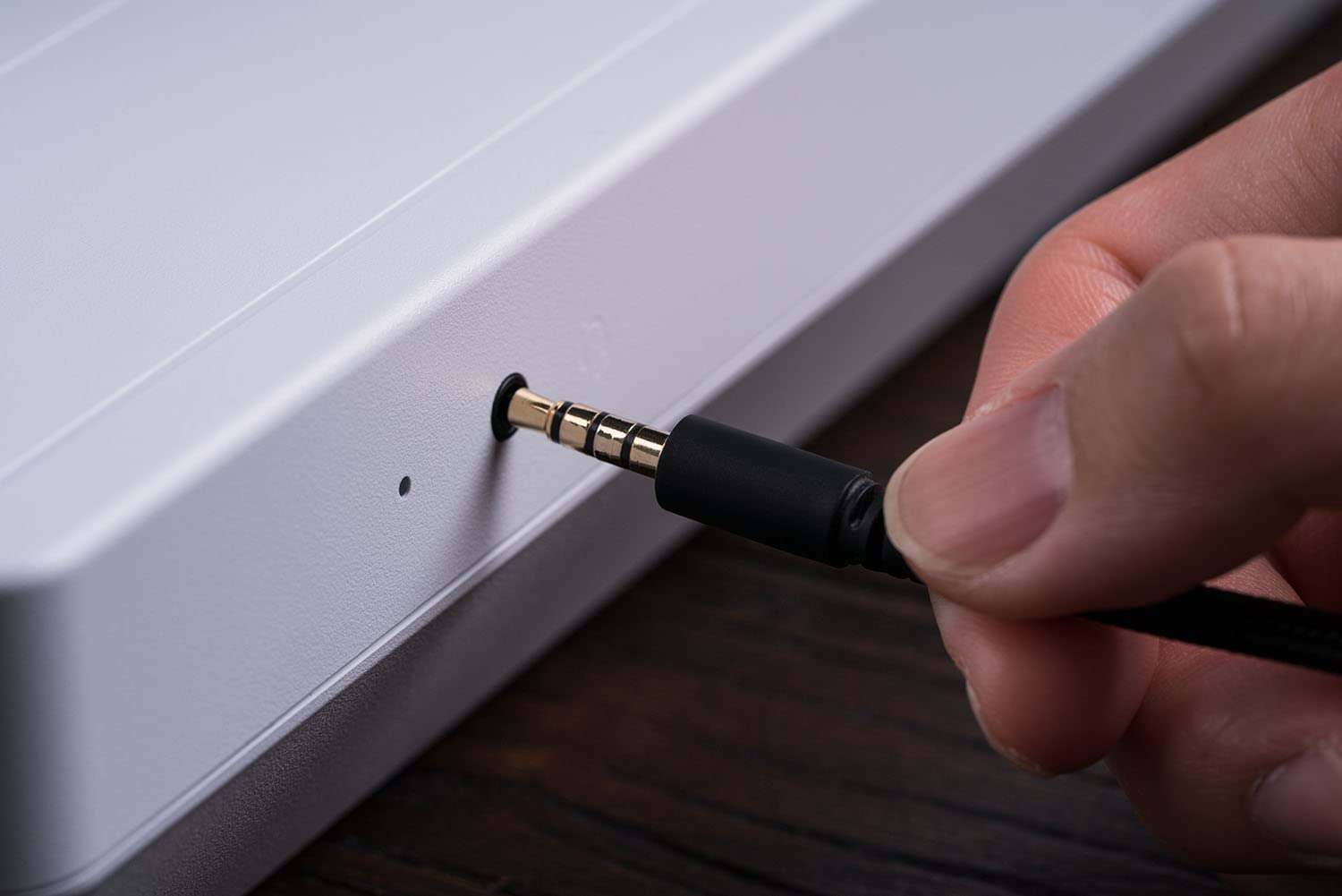 Ręka wkładająca czarny kabel do portu w szarym urządzeniu. Tło to drewno.