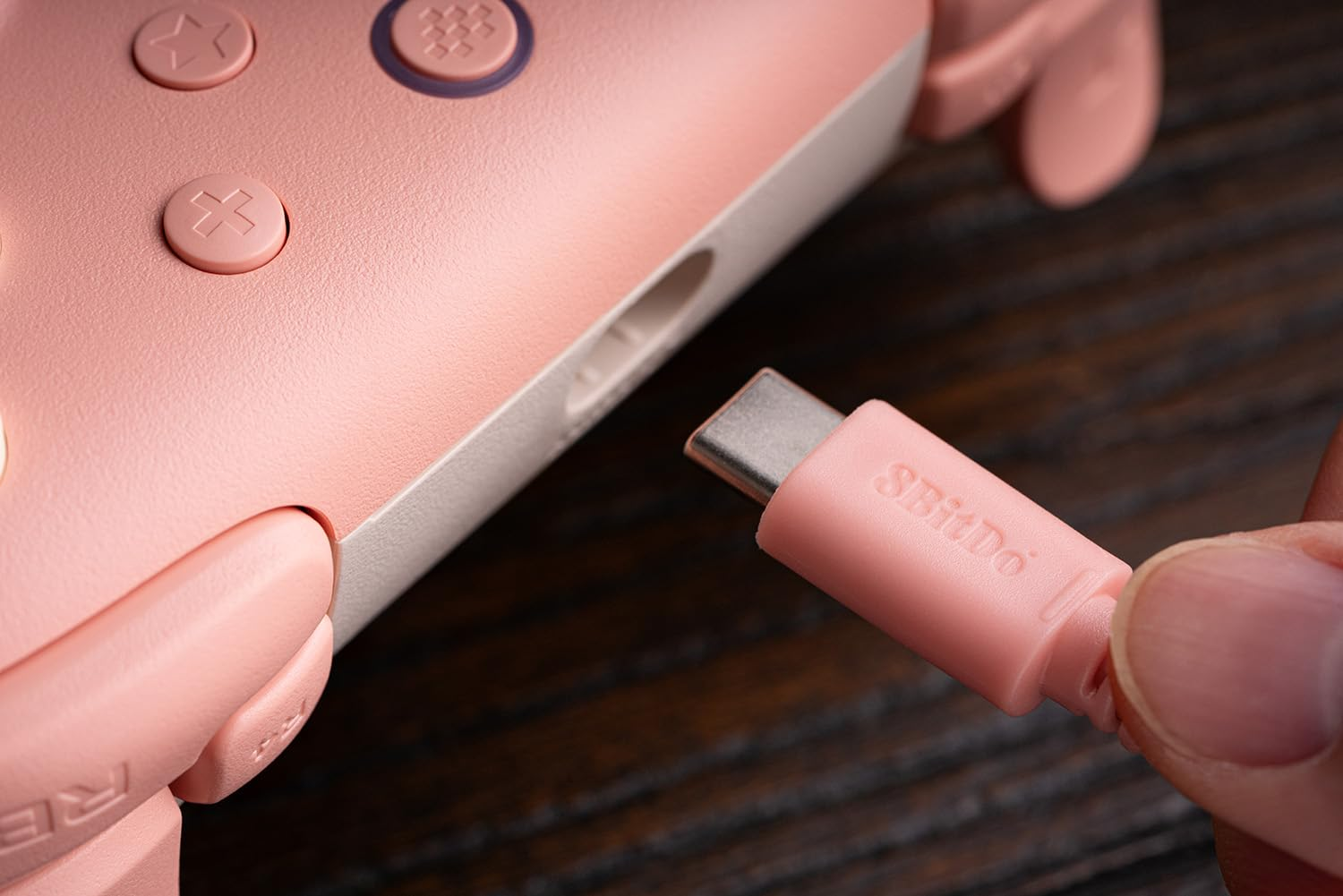 Zbliżenie różowego kontrolera 8BitDo podłączanego różowym kablem USB-C na drewnianej powierzchni.