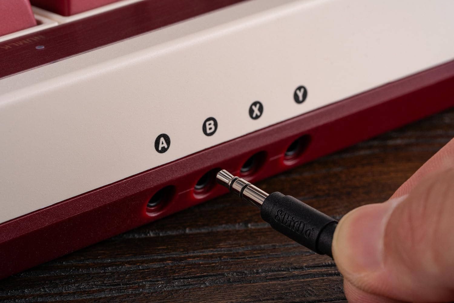 Ręka podłącza czarny kabel aux do klawiatury. Klawiatura ma czerwone i kremowe akcenty.