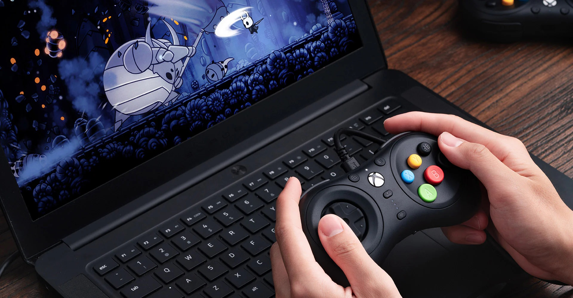 Mani che tengono un controller di gioco nero, che giocano su un laptop. Lo schermo mostra una scena di gioco.