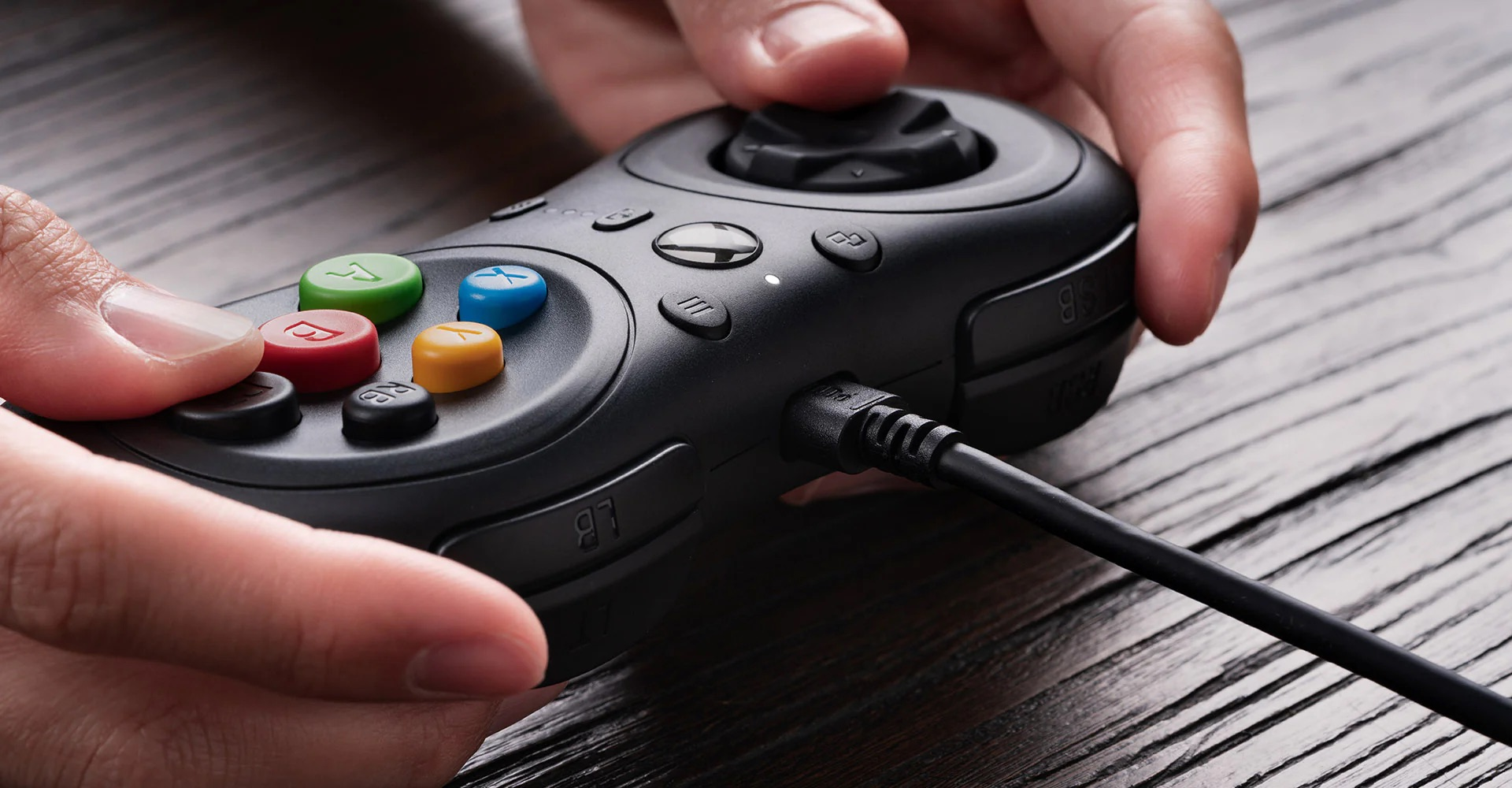 Primo piano di mani che tengono un controller nero, dita sui pulsanti. Tavolo di legno.