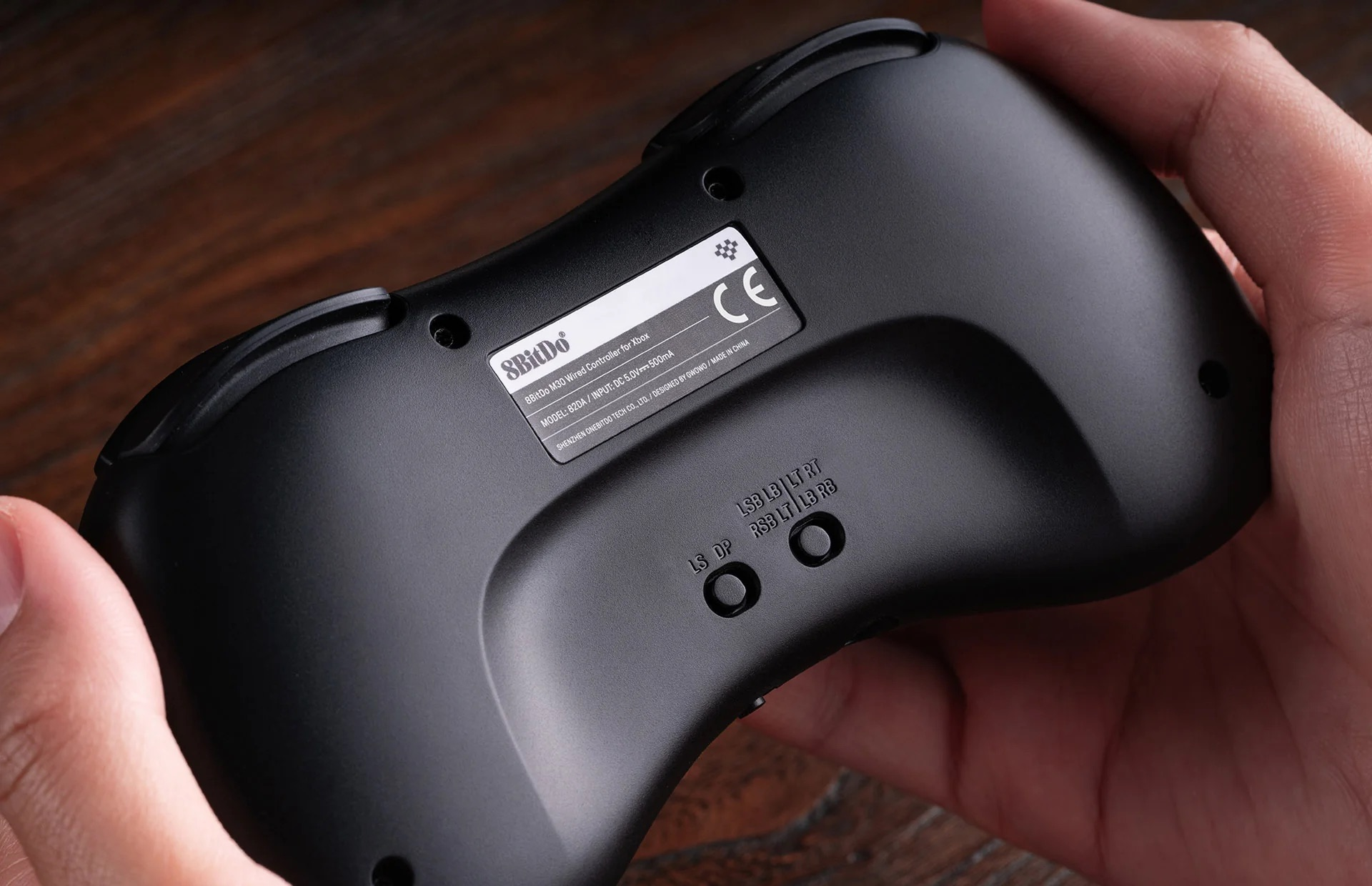 Primo piano del retro di un controller nero con etichette e un piccolo pannello. Tenuto da mani.