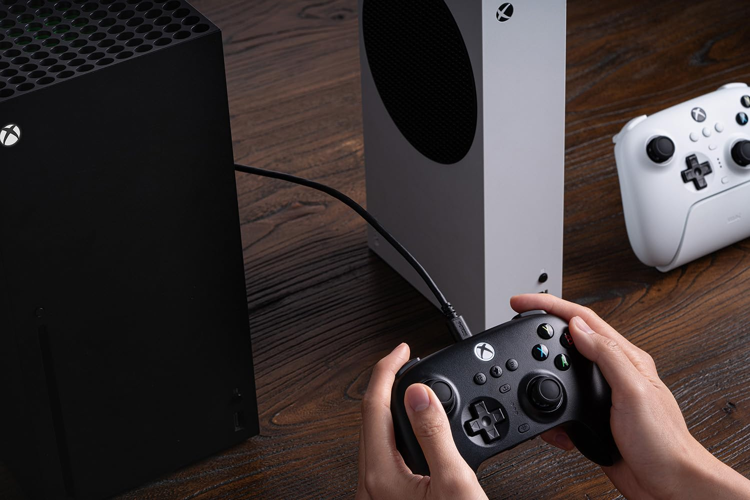 Mani che tengono un controller Xbox nero, collegato a console Xbox bianche e nere su un tavolo di legno.