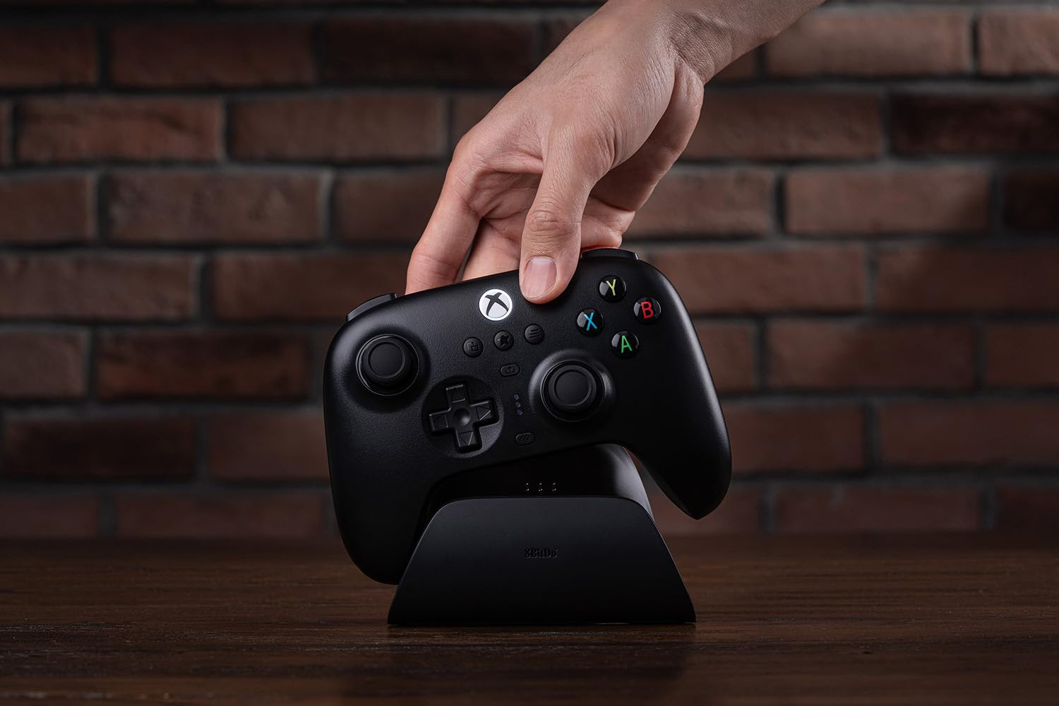 Una mano posiziona un controller Xbox nero sul suo supporto di ricarica. Lo sfondo è un muro di mattoni.