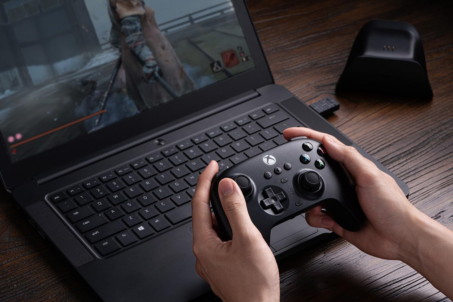 Mani che tengono un controller Xbox, che giocano a un gioco su un laptop, con un supporto di ricarica sullo sfondo.