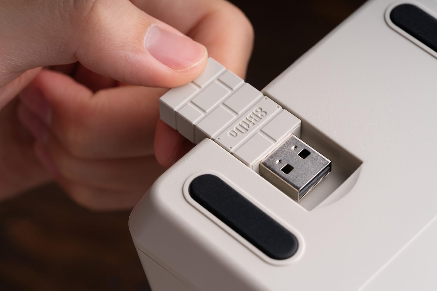 Una mano che inserisce un mattone USB grigio in una tastiera. L'USB è marchiato '8BitDo'.