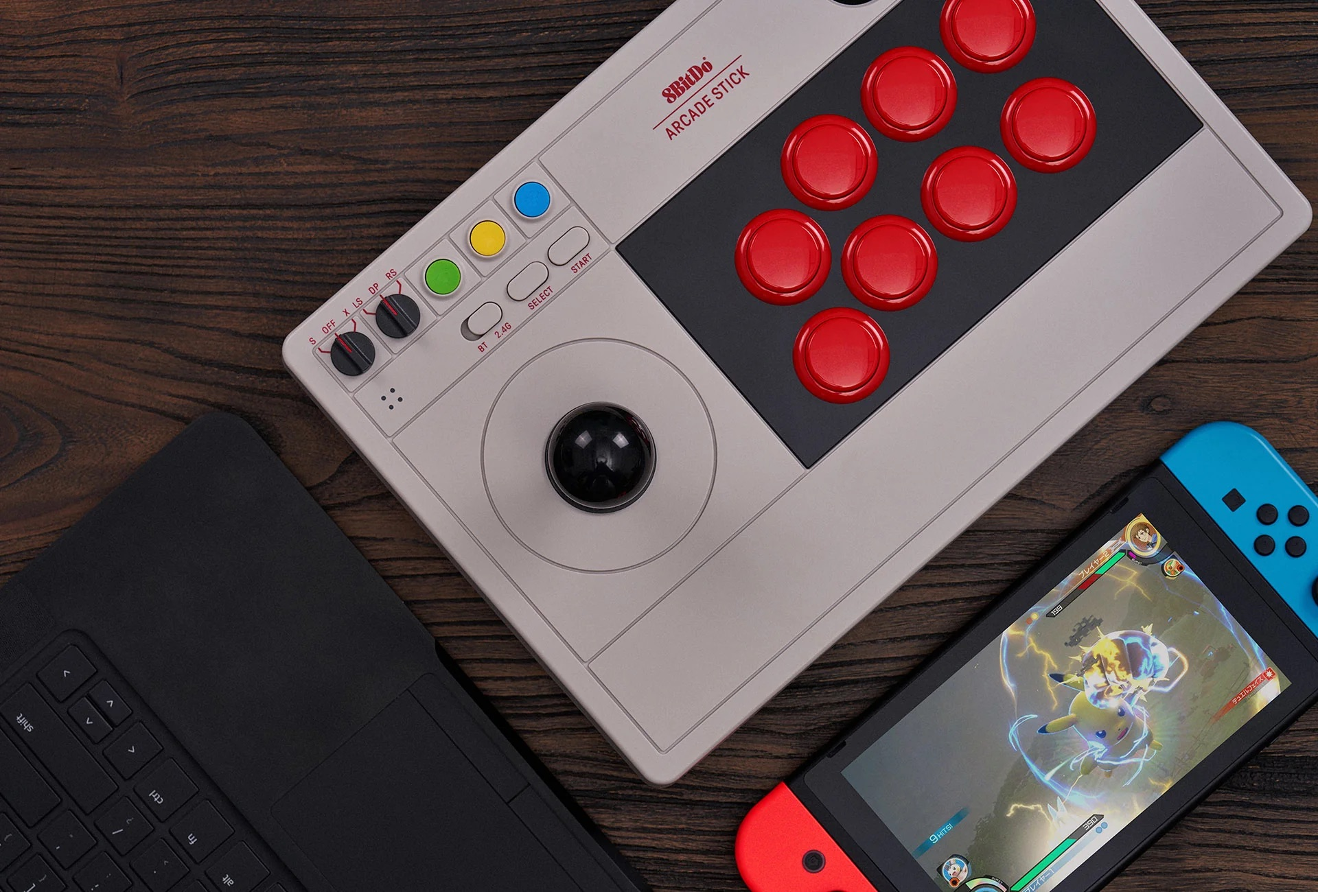 Un arcade stick 8BitDo con joystick nero, pulsanti rossi, e una Nintendo Switch e un laptop vicino.