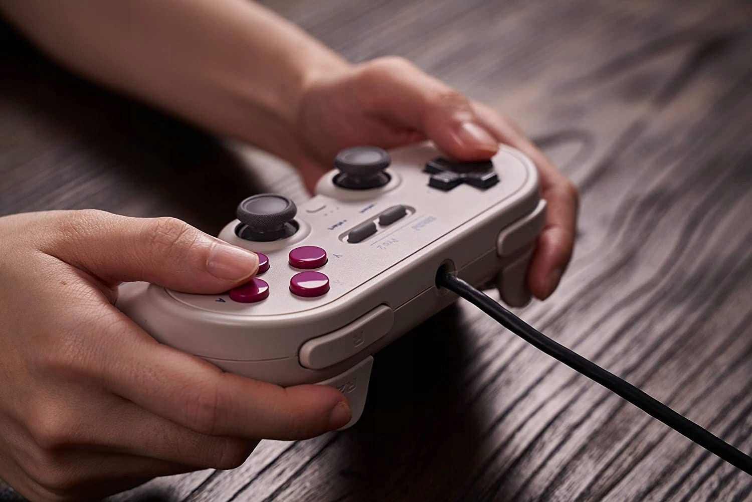 Dłonie trzymające biały gamepad z szarymi joystickami i różowymi przyciskami na drewnianej powierzchni.