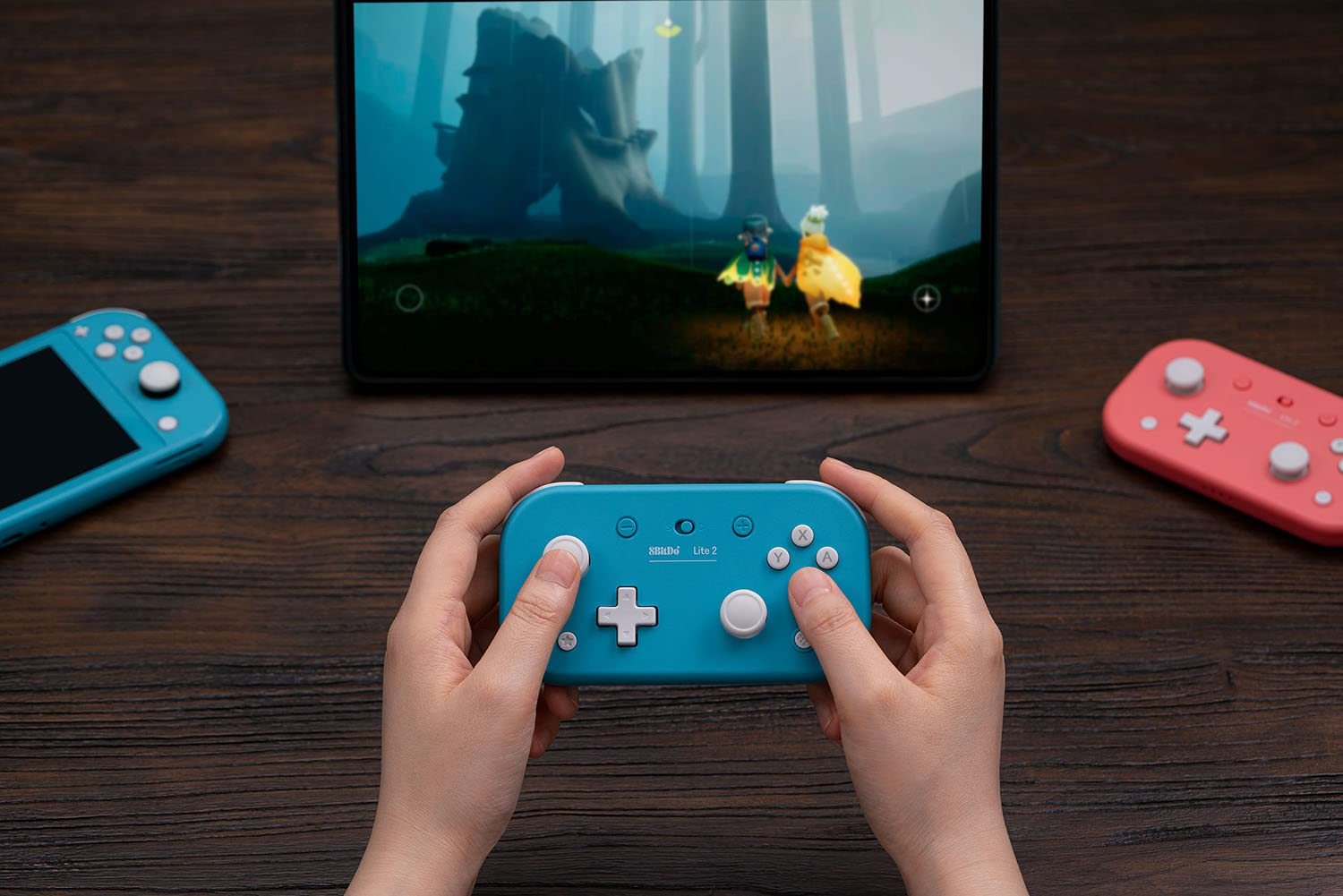 Ręce trzymające niebieski gamepad, z tabletem pokazującym grę i różowy gamepad.