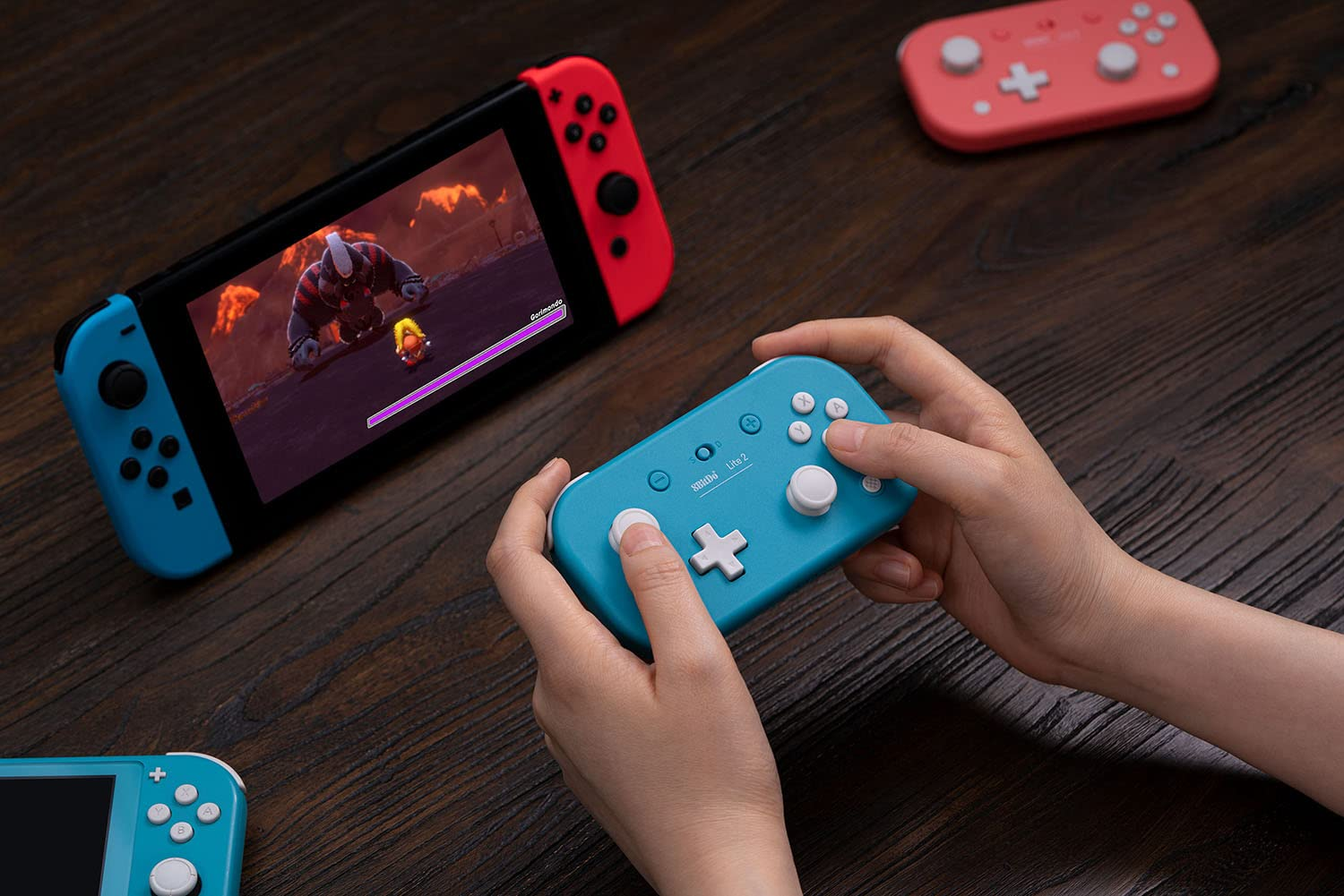 Dłonie trzymające niebieski kontroler, Nintendo Switch i różowy kontroler na drewnie.
