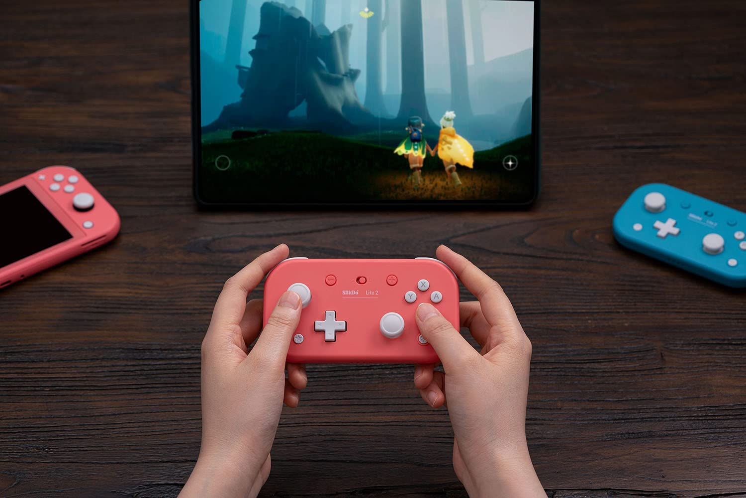 Mani che tengono un controller rosa, un tablet che mostra un gioco e un controller blu su una superficie di legno.