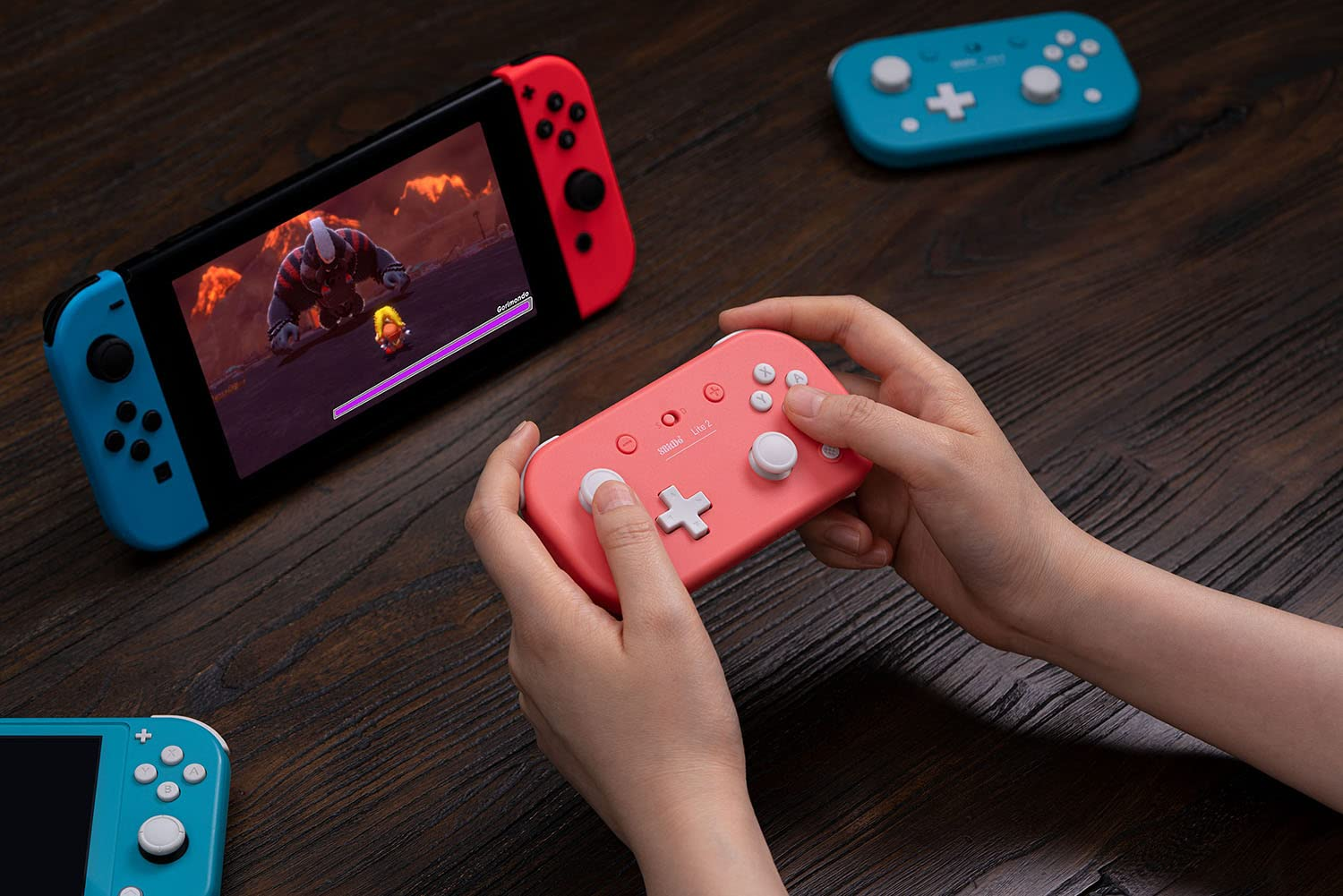 Mani che tengono un controller rosa, Nintendo Switch e un controller blu su una superficie di legno.
