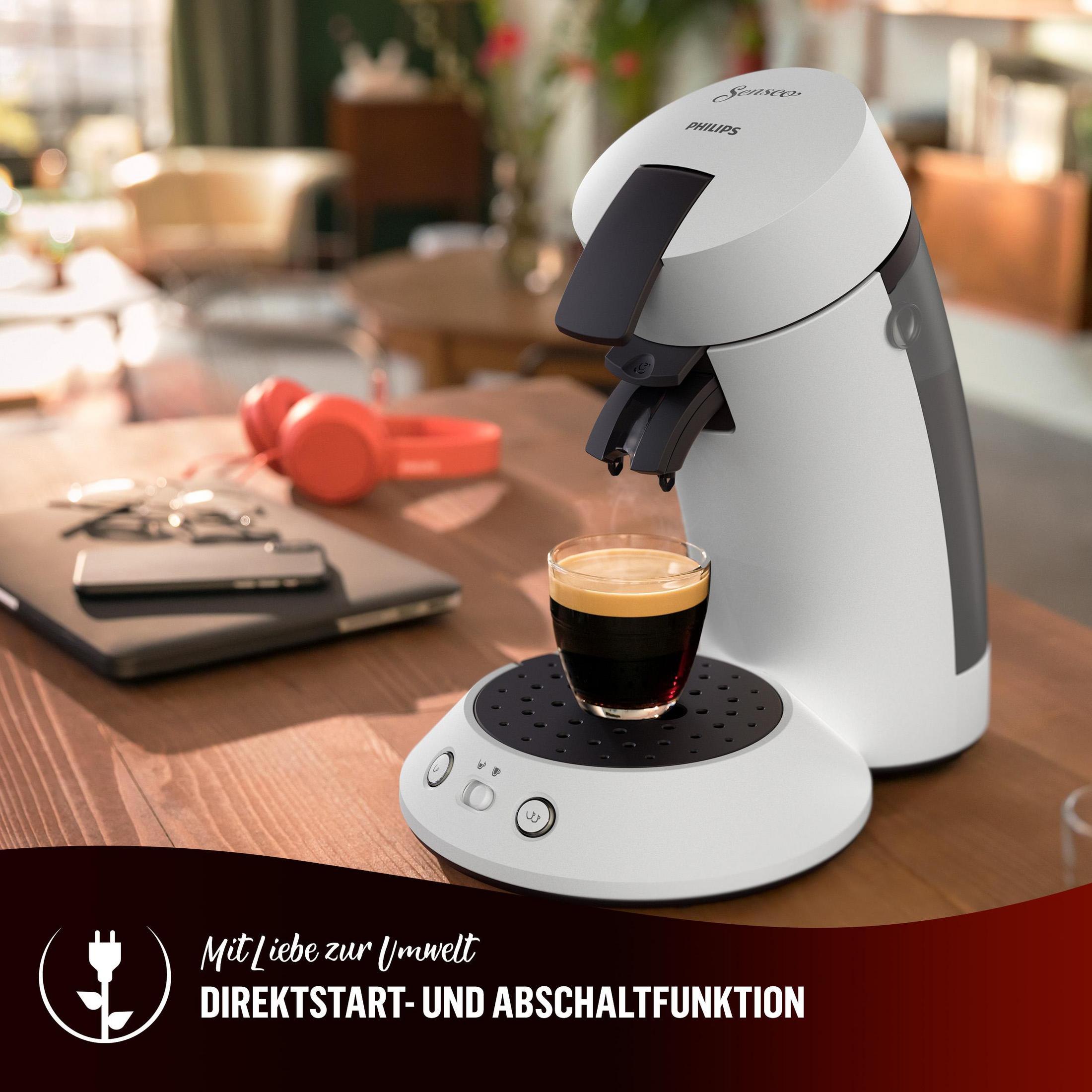 Weiße Kaffeemaschine, die Kaffee in ein Glas brüht. Kopfhörer und Laptop auf einem Tisch.