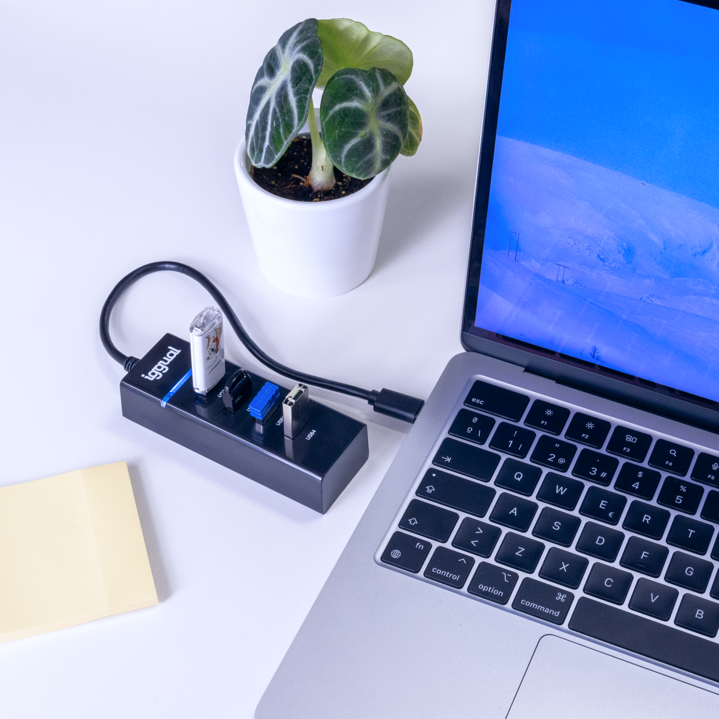 Laptop con un concentrador USB, una planta y una nota adhesiva amarilla sobre un escritorio blanco.