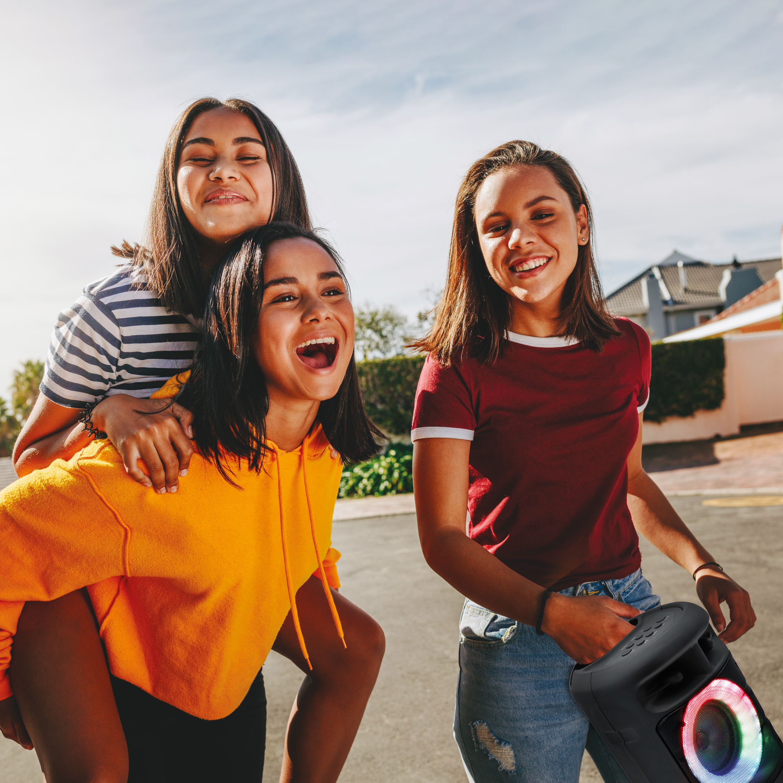 Tres mujeres jóvenes sonriendo y riendo. Una es llevada a caballito. Hay un altavoz en la escena.