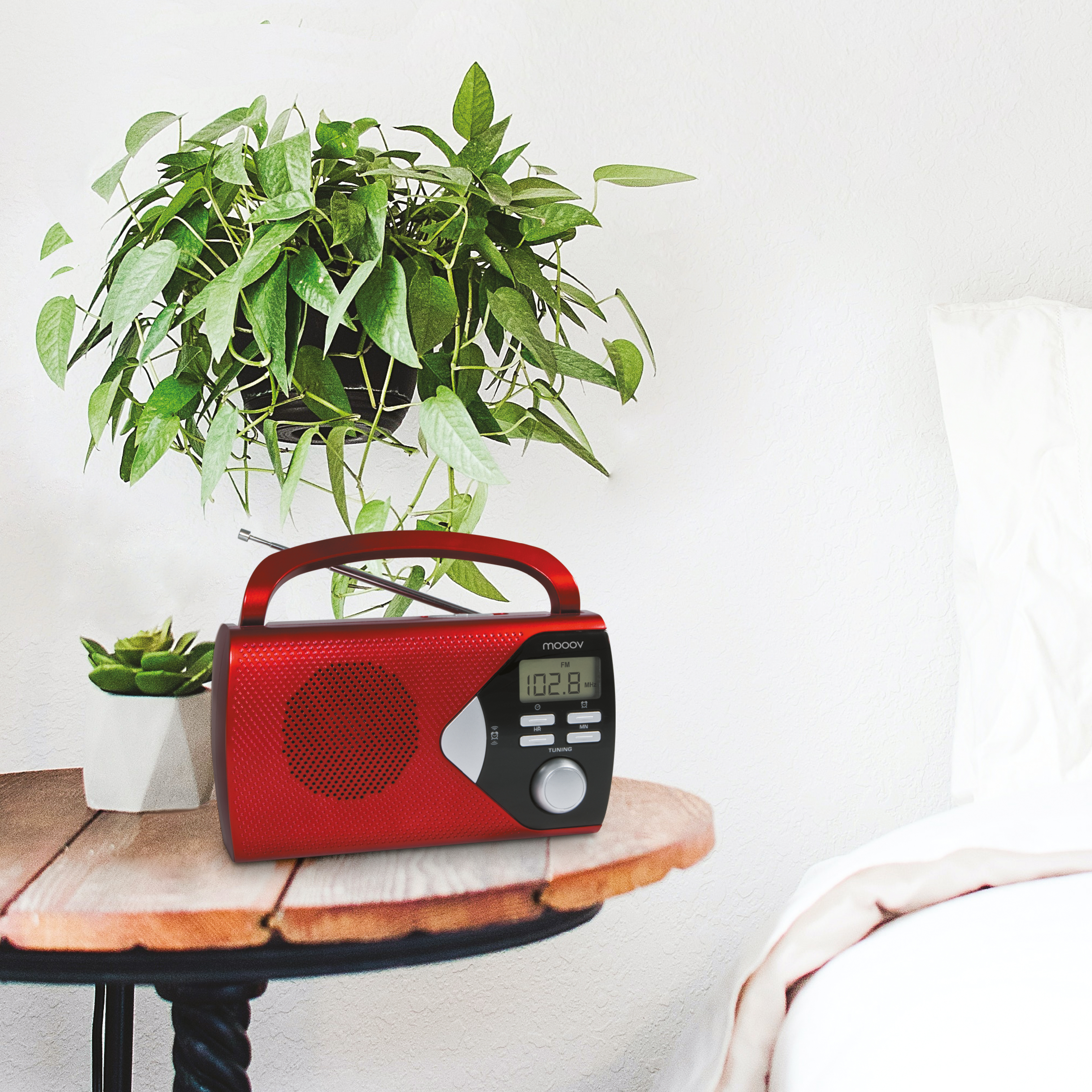 Radio rojo sobre una mesa de madera junto a una cama blanca. Una planta está encima de la radio.