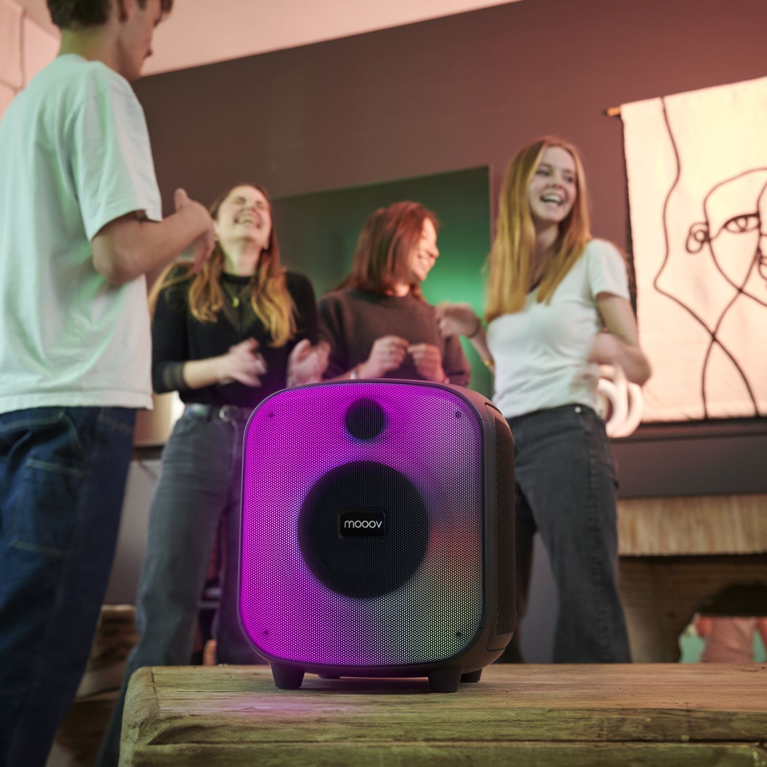 Un altavoz en una mesa de madera. Cuatro personas en el fondo, algunos riendo y bailando.