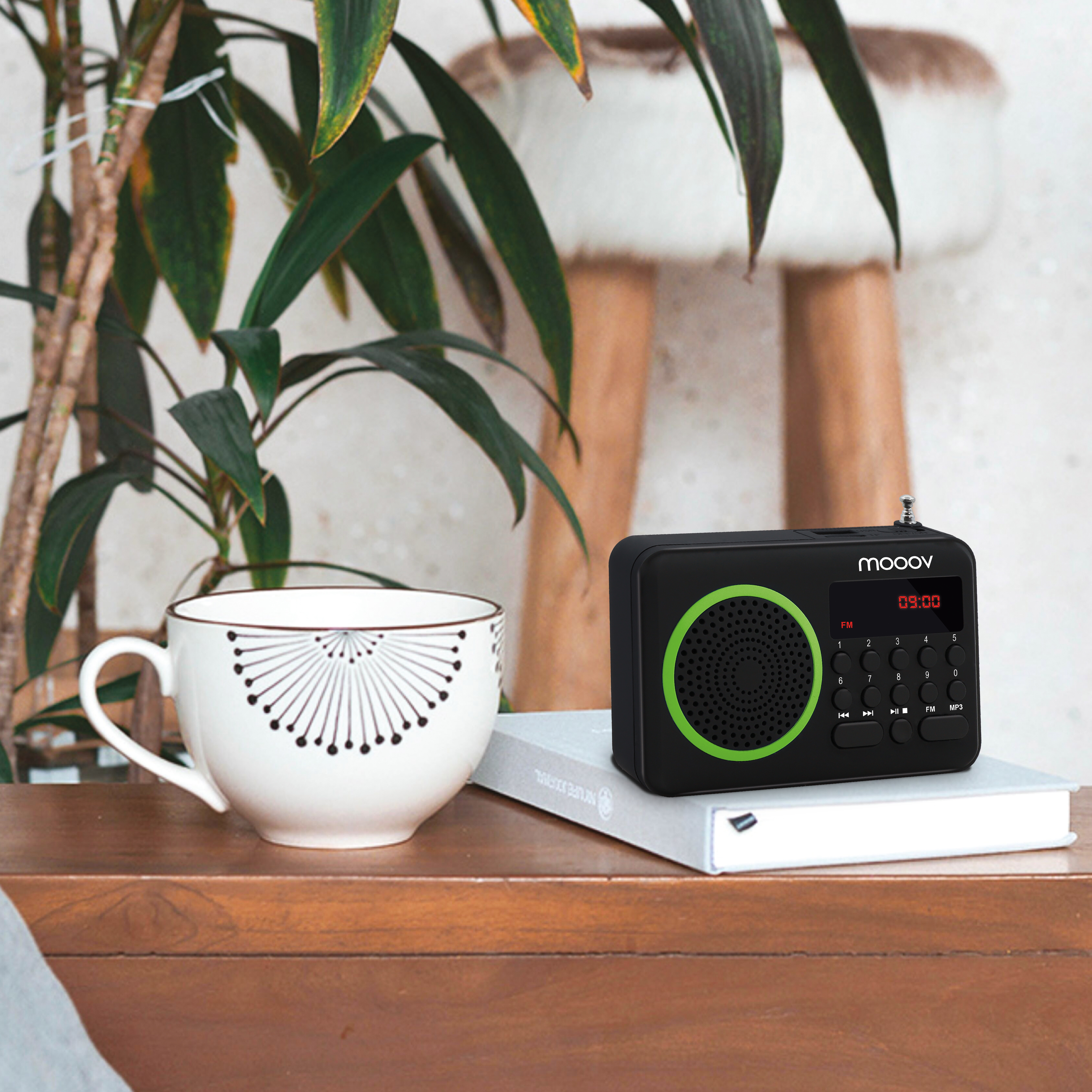 Radio negro con anillo verde, sobre libro blanco, junto a taza y planta.